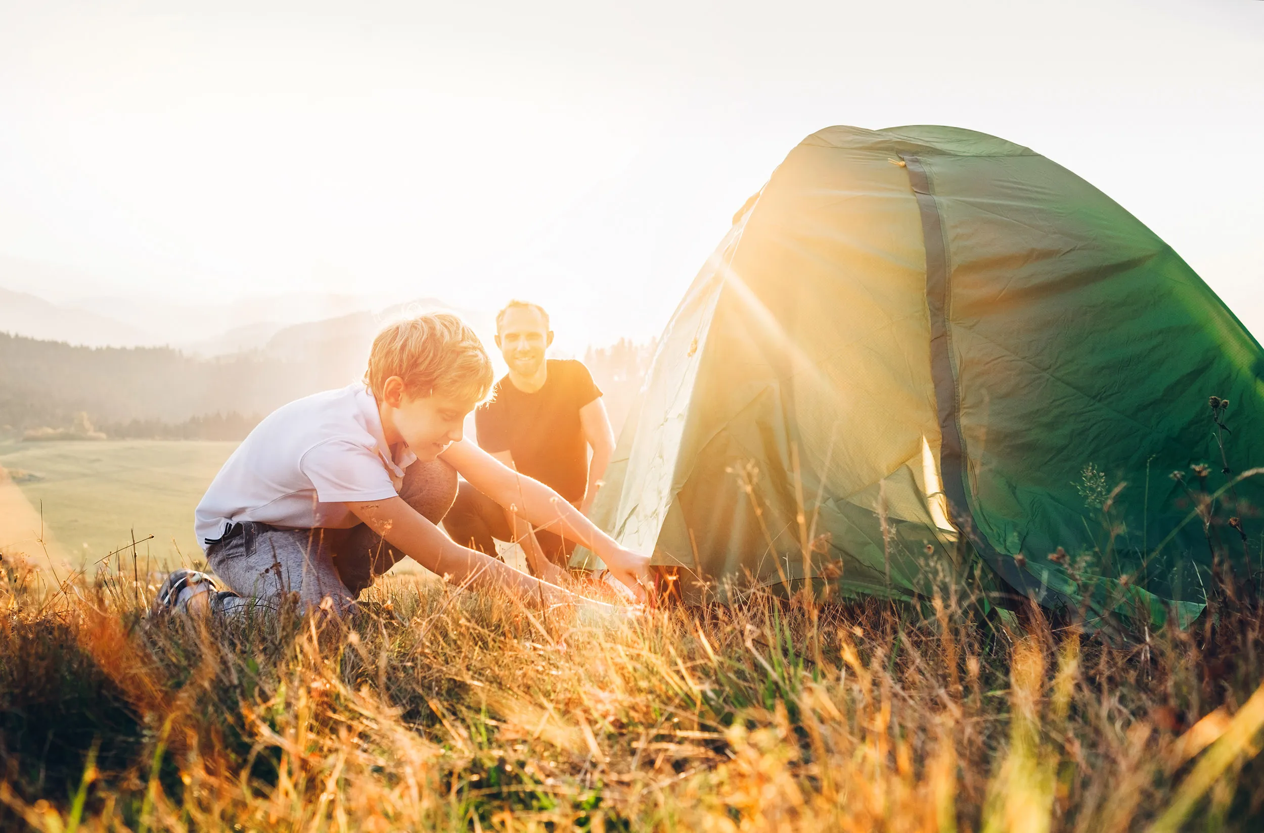 An adult and child setting up a tent on a grassy hill, with the sun sunsetting behind.