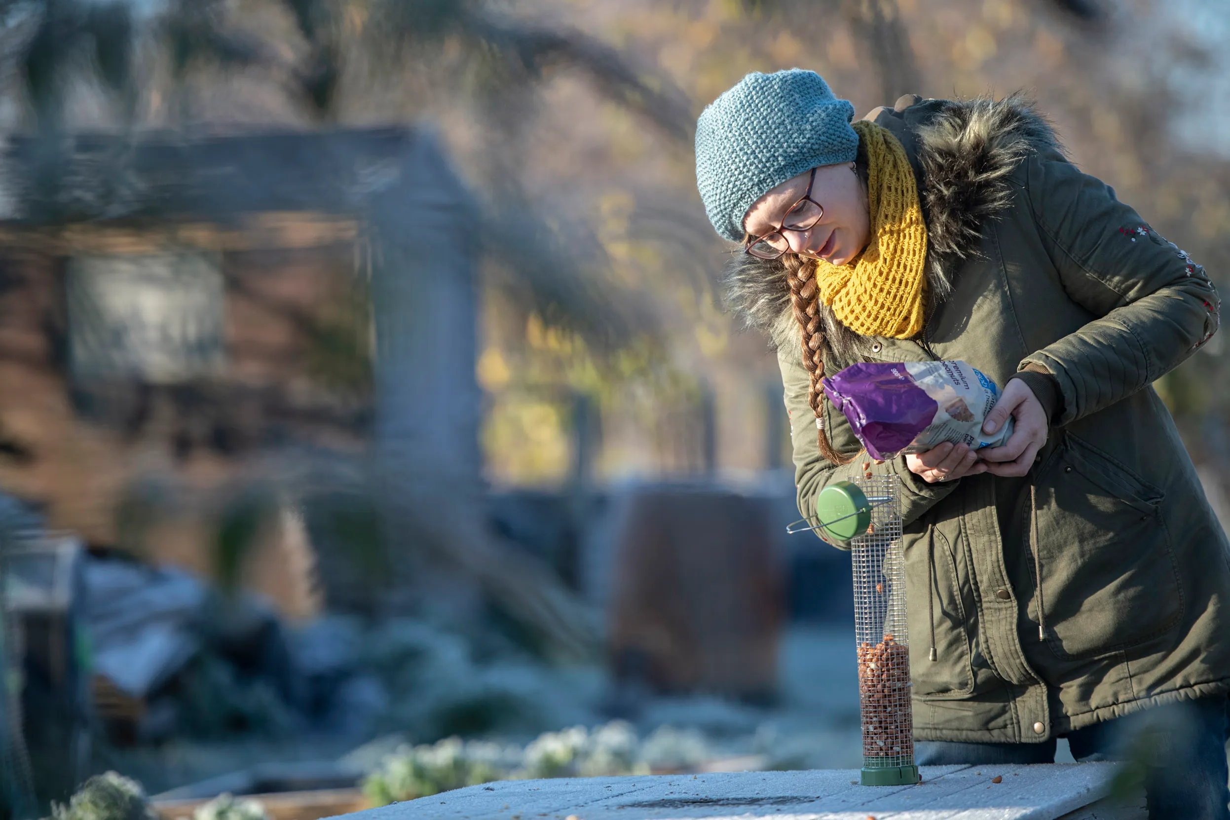 A person wearing a hat, coat and scarf filling a bird feeder on a table outside during winter.