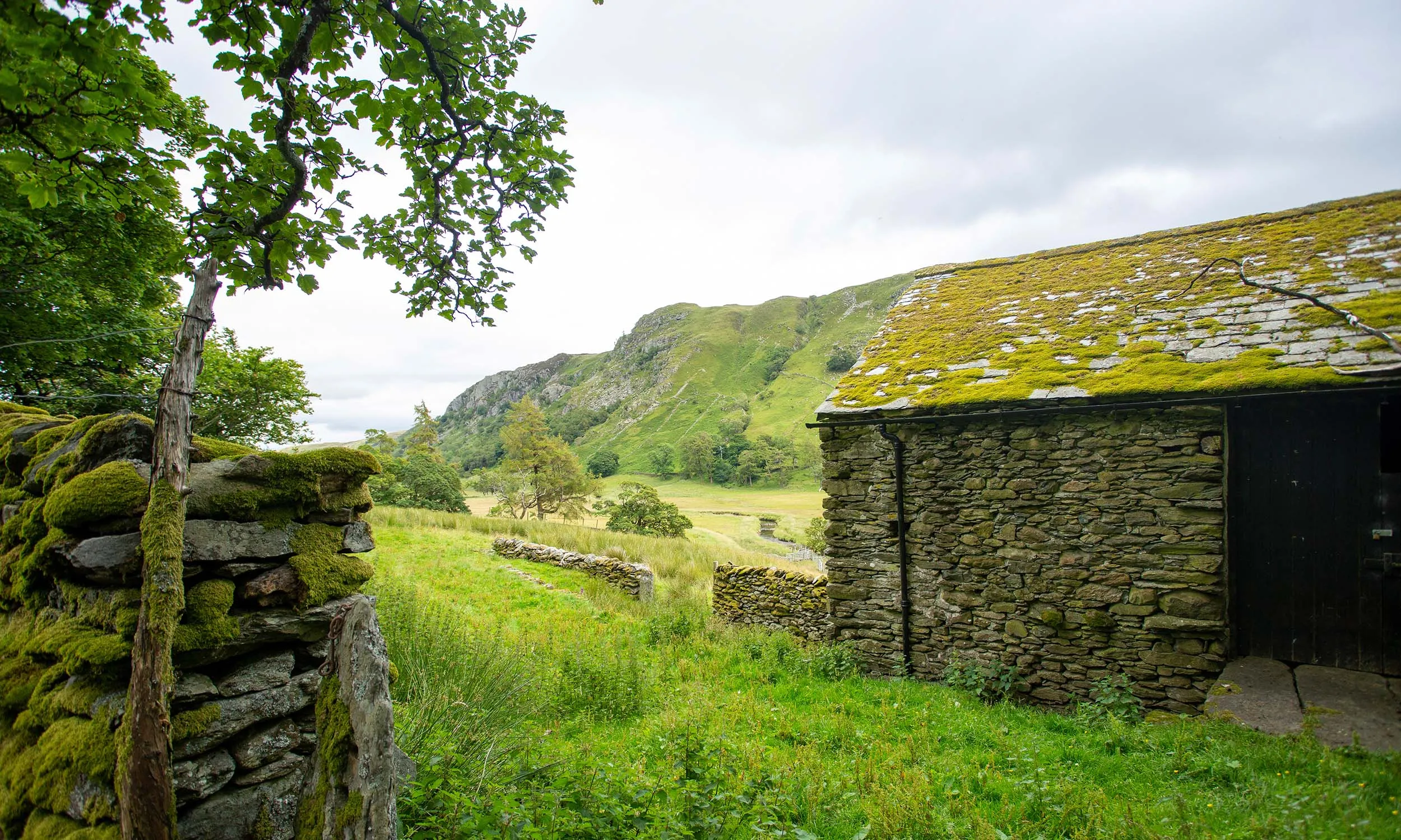 A mossy stone and slate farm building, stood on a grassy plot of land with a steep hill along the horizon.