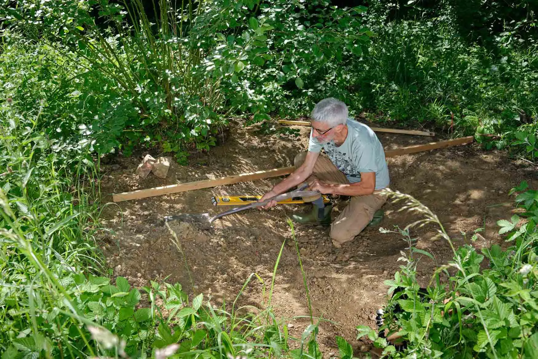 A man crouched down in amongst tall grass digging out a large pond.