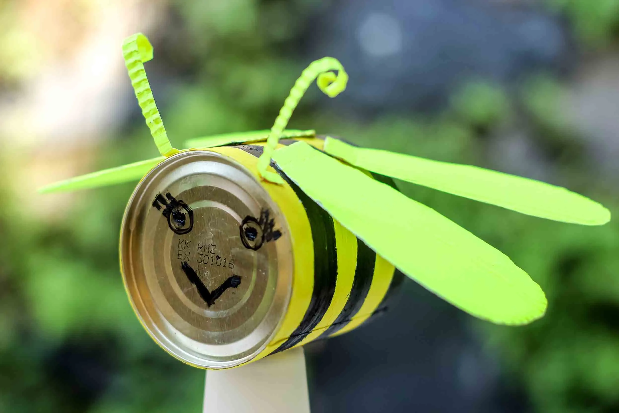 A tin can fashioned to resemble a bee by the addition of paper wings and antenna, the colouring of black and yellow stripes around the curved side of the can, and a face drawn onto the bottom surface.