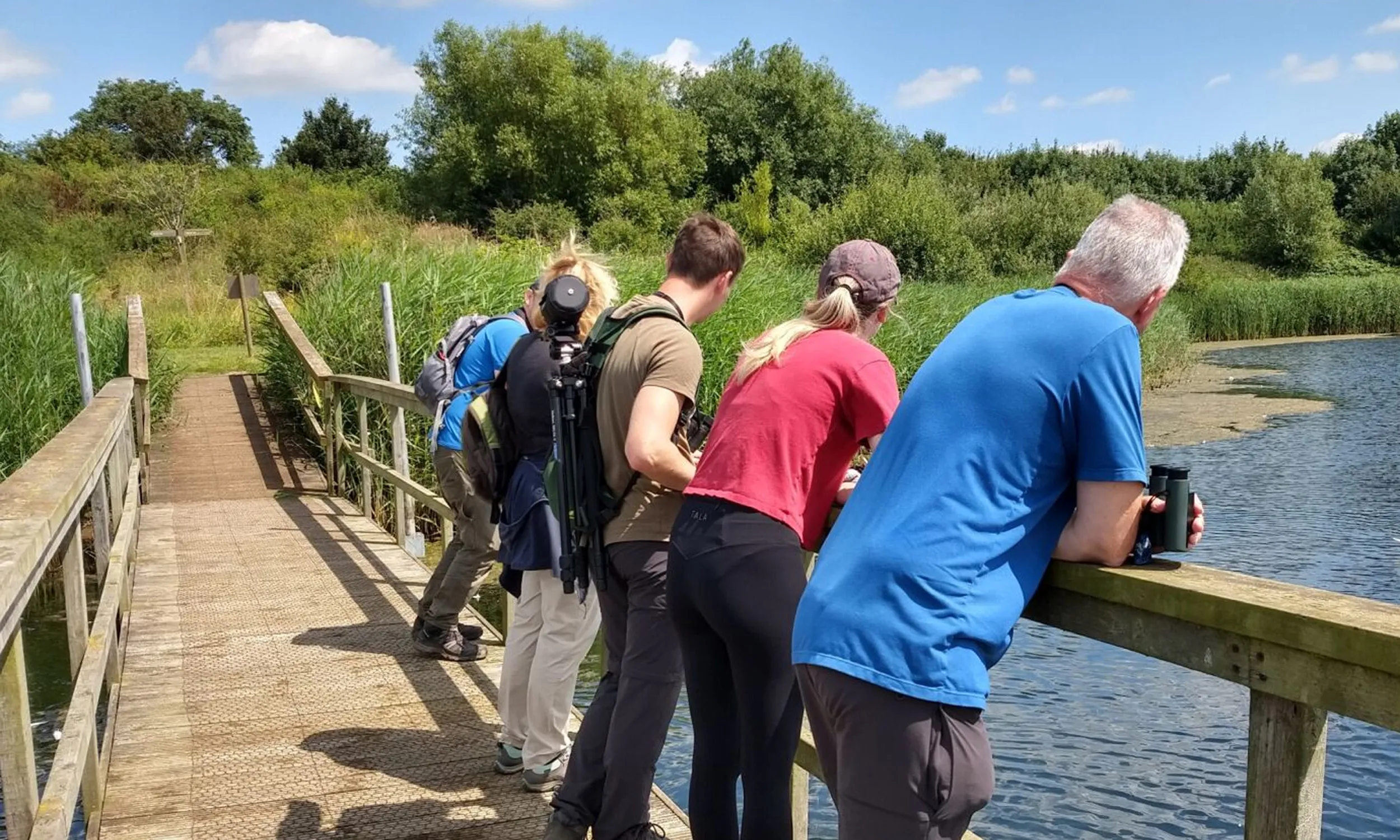 People leaning on the wooden handrail of a bridge that crosses a body of water at Langford Lowfields