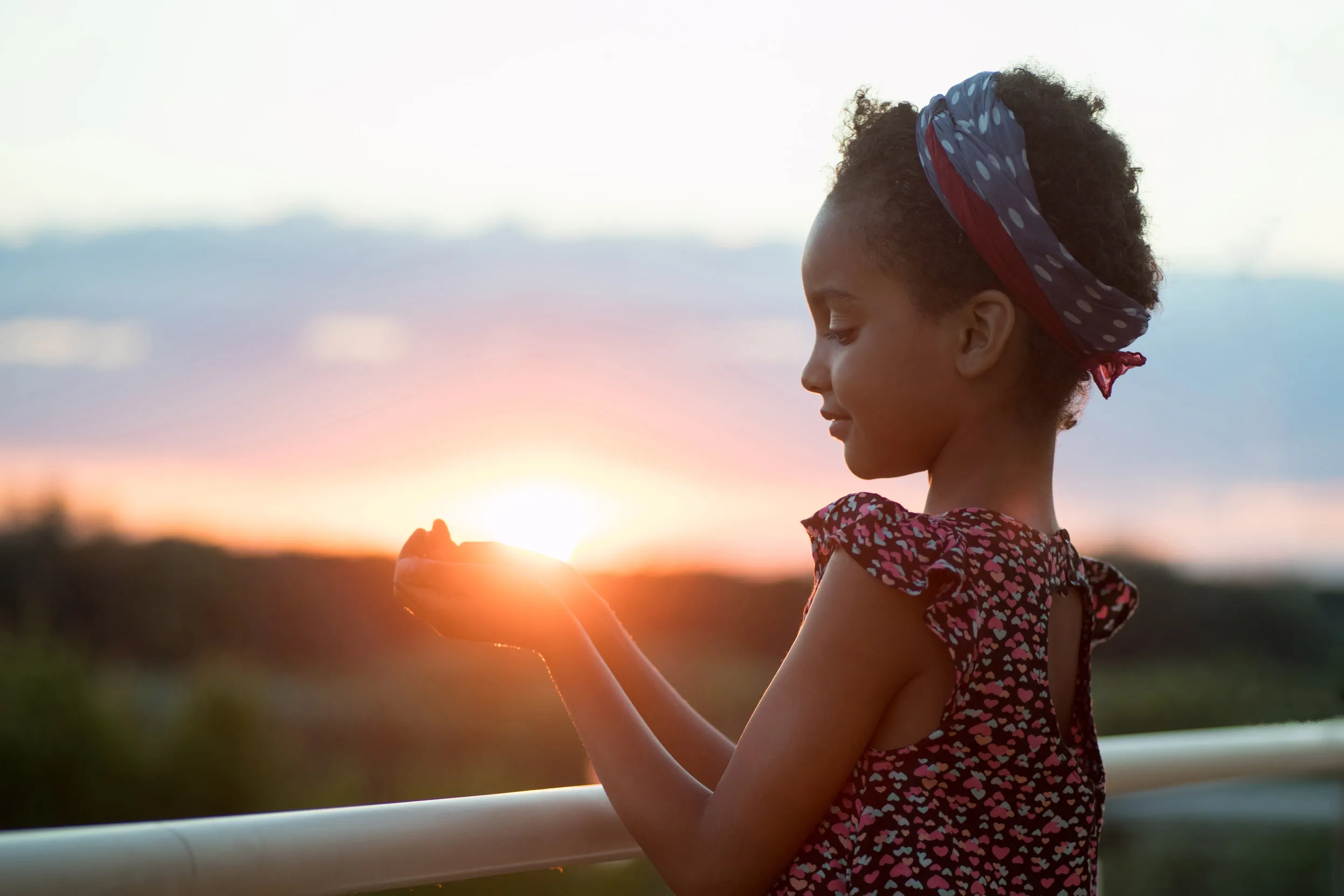 The side view of a child cupping the sunset in their hands stood on a bridge. 