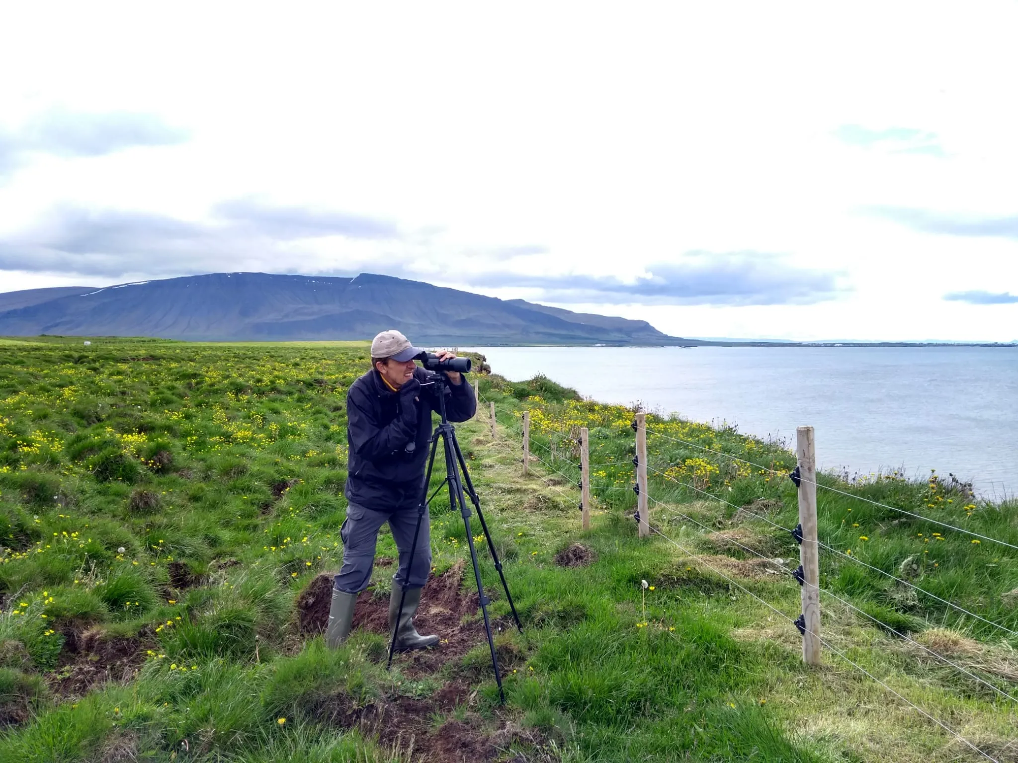 A researcher wearing a cap looks through a monoscope standing in a field in front of a fence, by the sea.