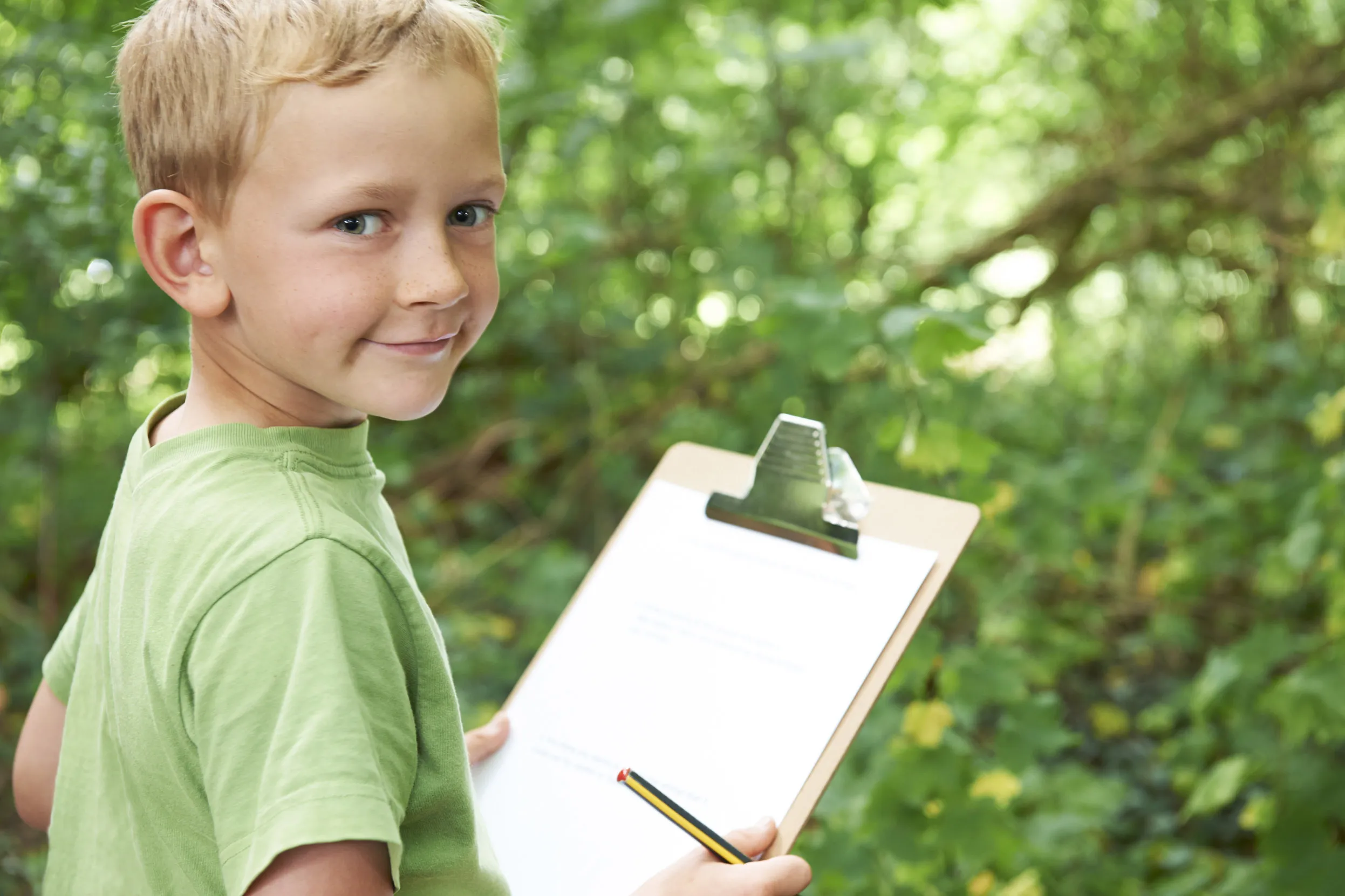 A child wearing a green top writing notes on a clipboard in the woods.
