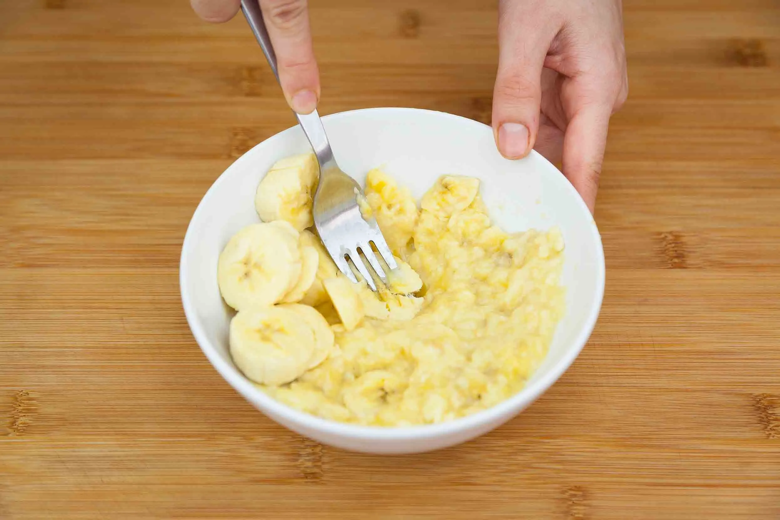 A person's hands mashing a sliced banana in a bowl with a fork.