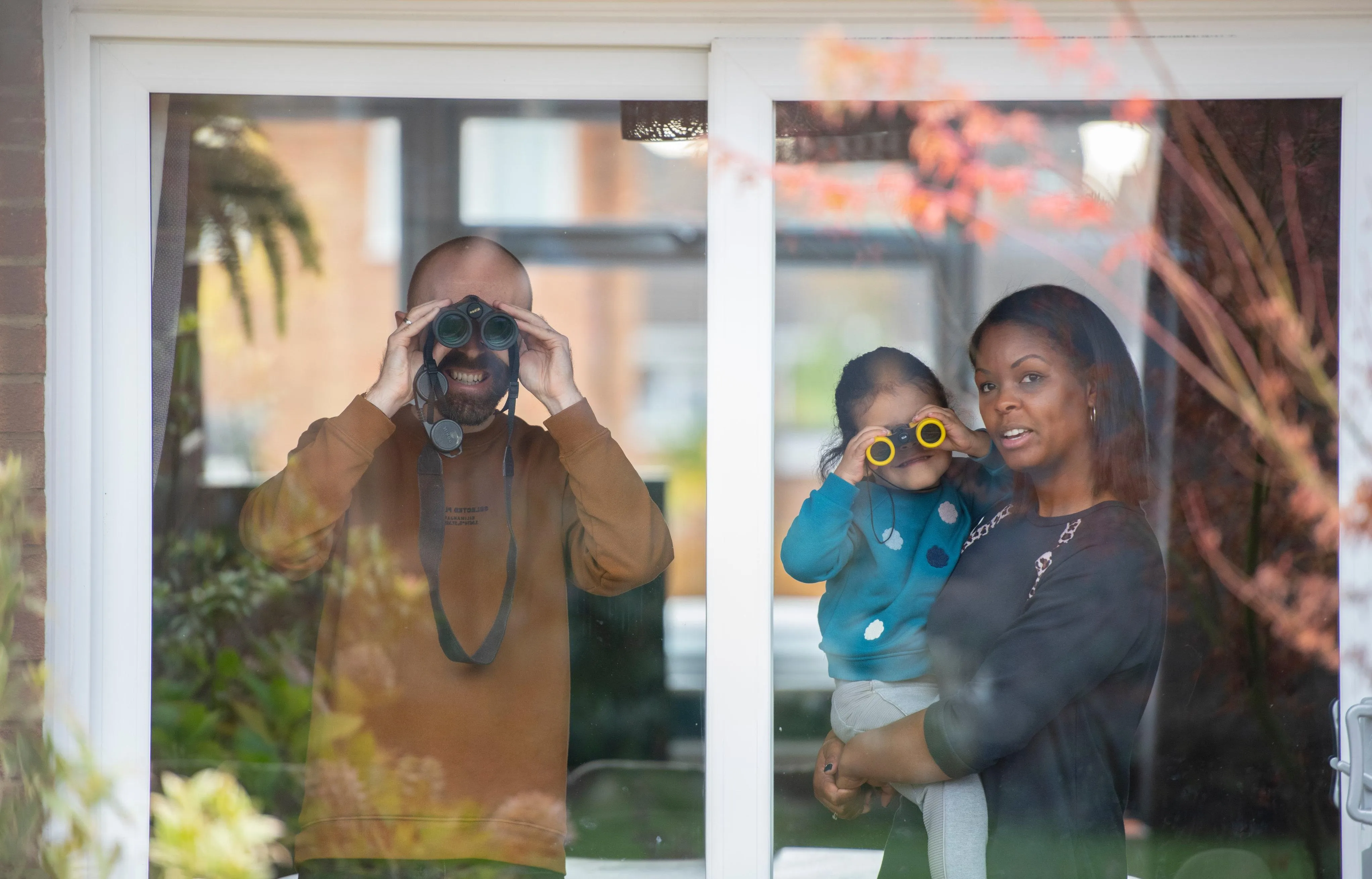 Two adults and a child stood behind a glass door looking out into the garden, one of the adults and the child look through pairs of binoculars.