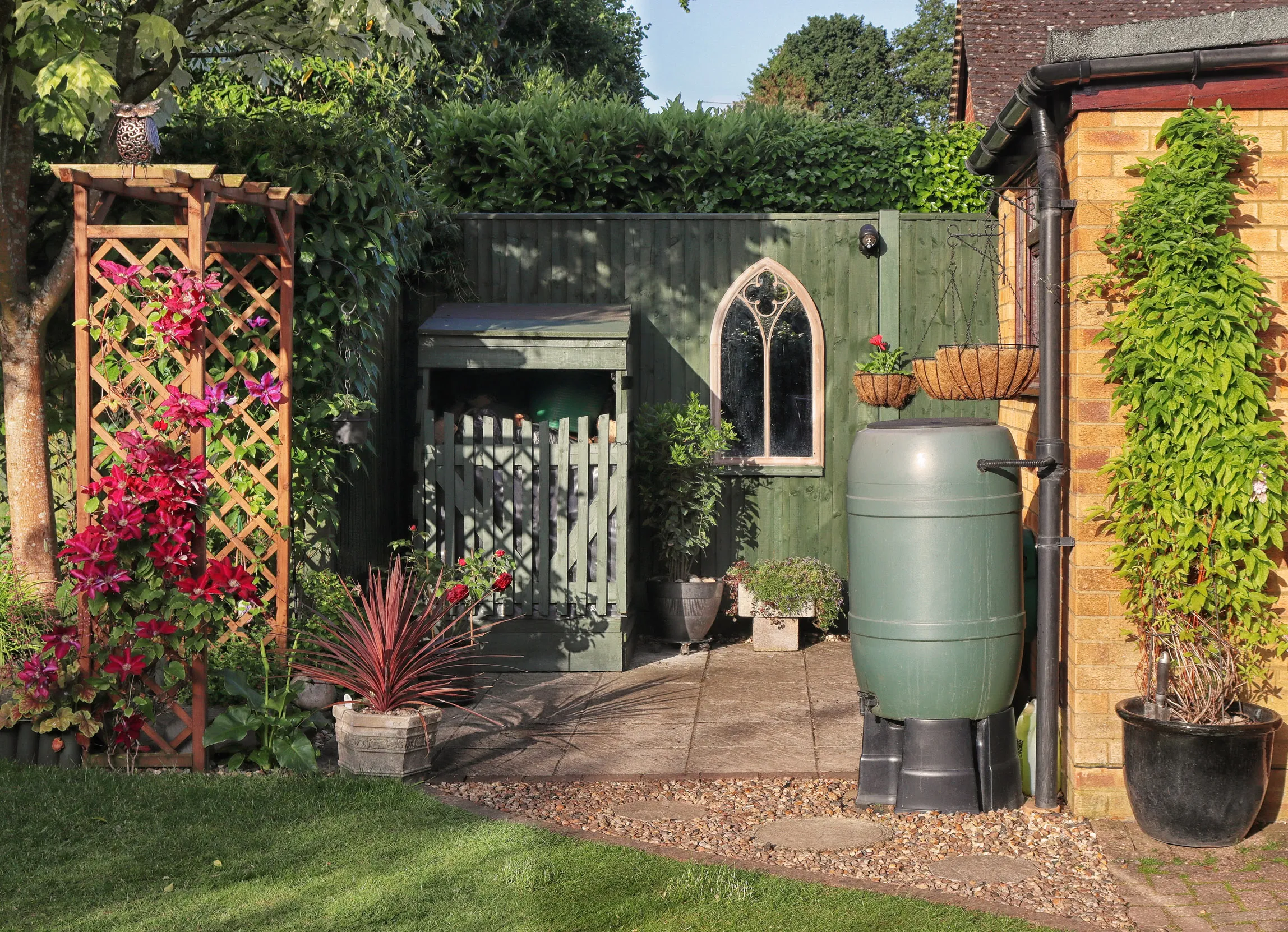 The corner of a sunny garden, featuring a small patio, lawn, a plastic water butt and a small lean-to wood store.