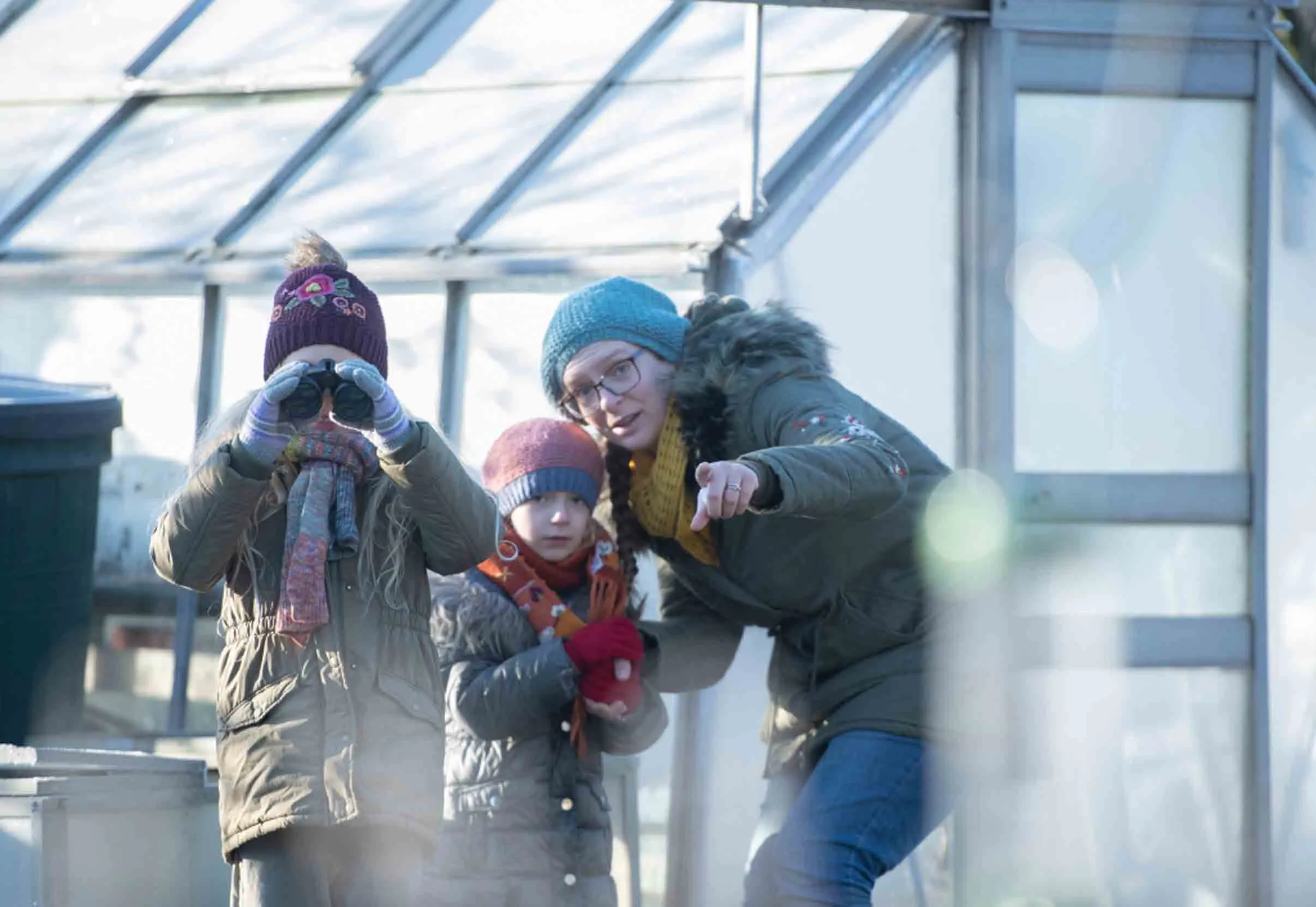 An adult and two children wearing warm winter clothing in front of a frosty greenhouse, one child is looking through binoculars and the other is looking to where the adult is pointing.