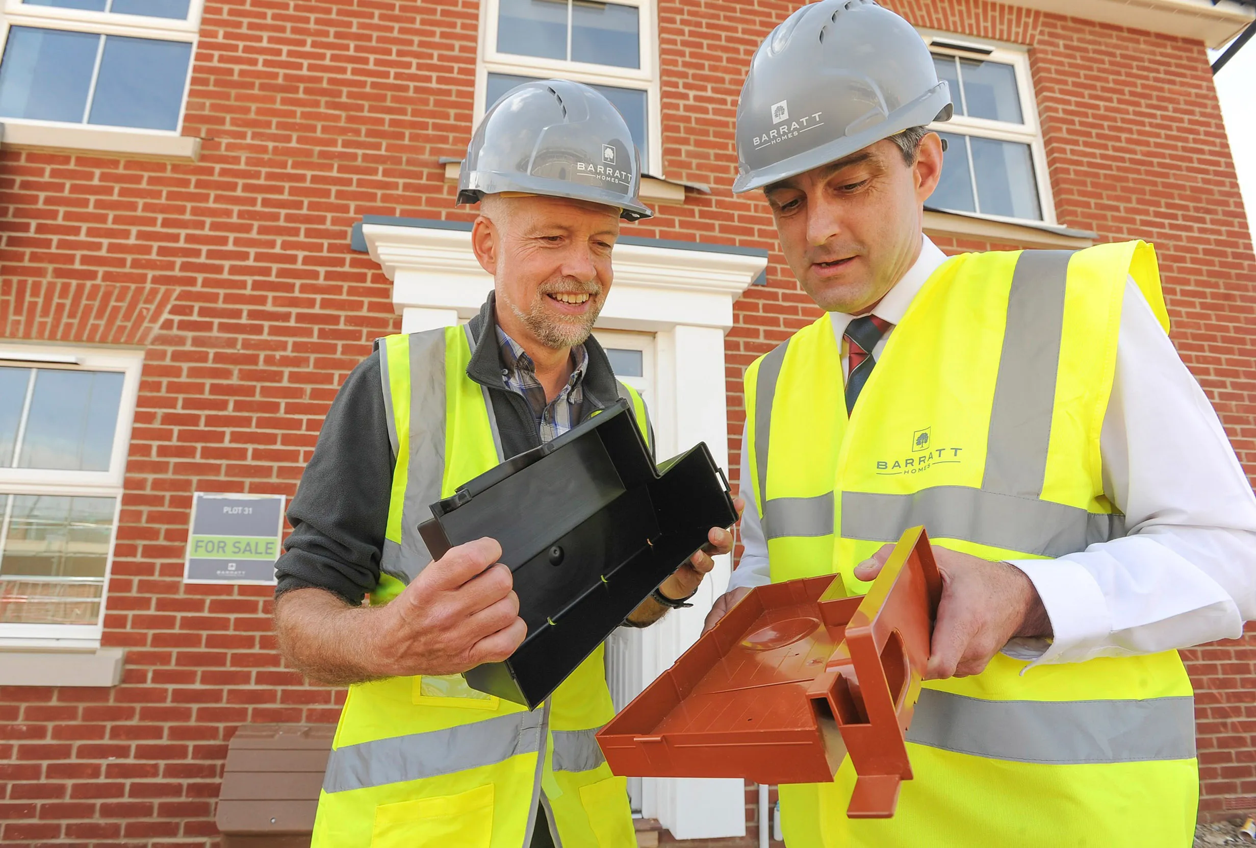 Two men, with hard hats and hi vis on, stood outside of a new build Barratt home holding a swift brick.