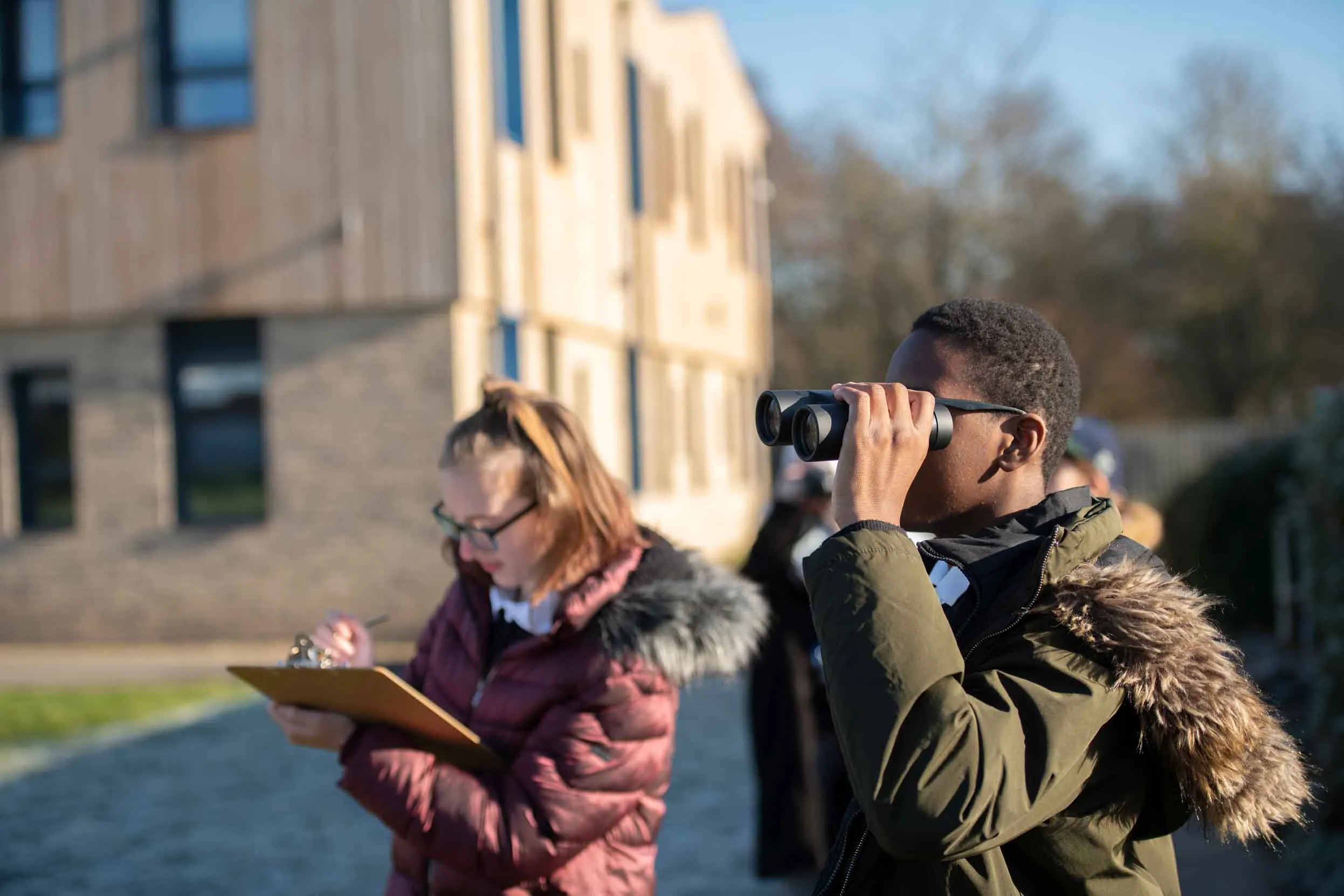Two children outside of their school building, one in a red coat writing on a clipboard and the other in a green coat looking through binoculars.