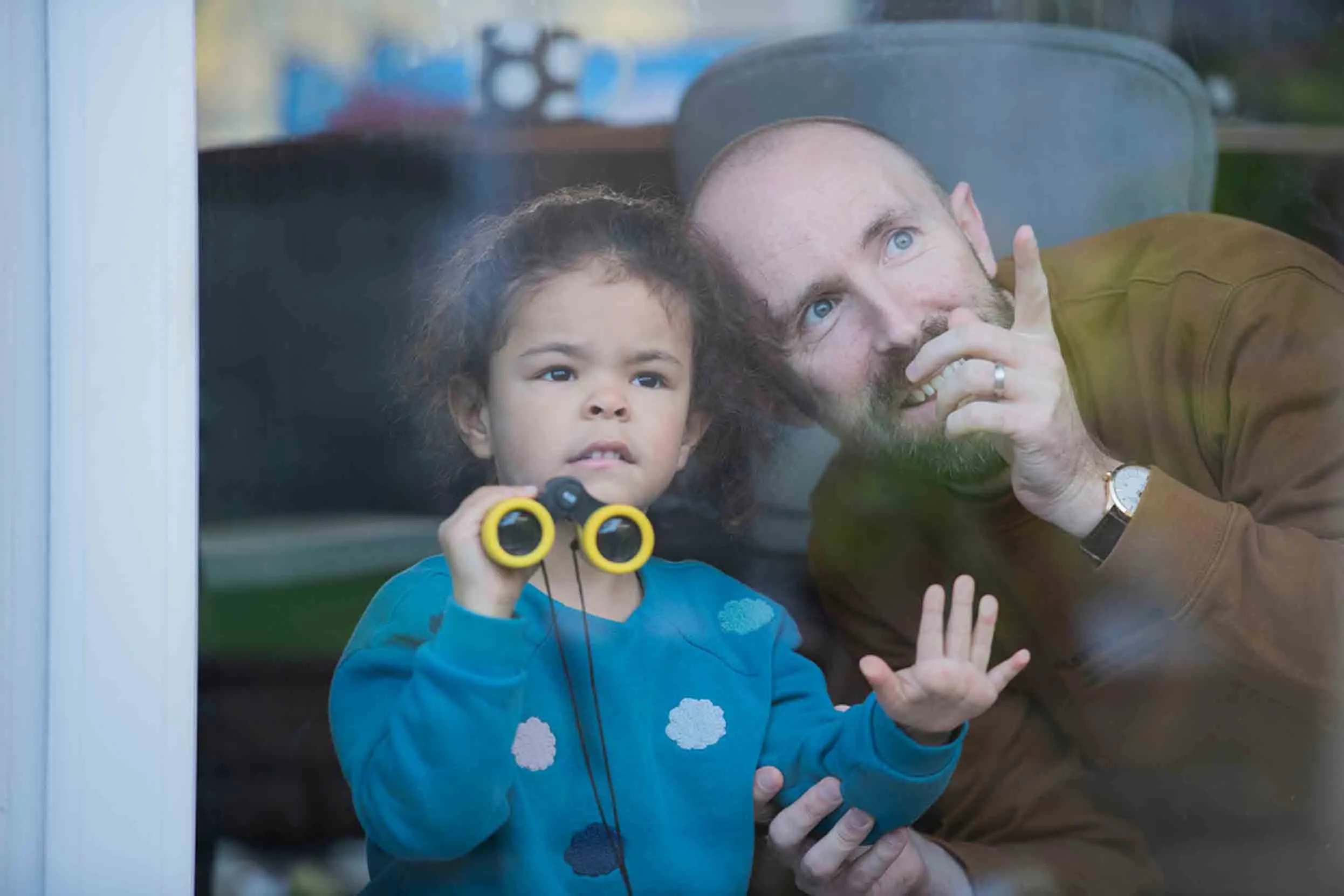 An adult and a small child birdwatching from inside a house, behind a large window. The child is holding yellow binoculars and the adult is smiling and pointing to something outside.