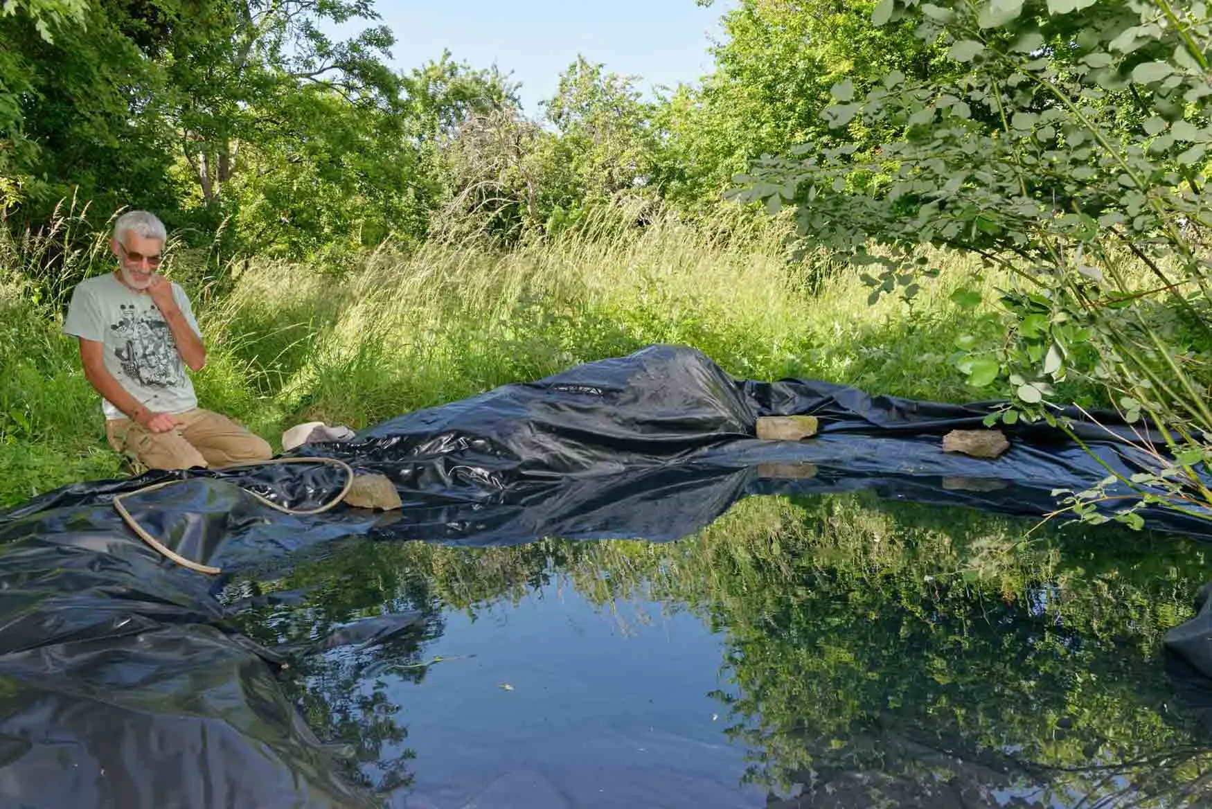 A man sat beside his large pond in enjoyment as he fills it up.