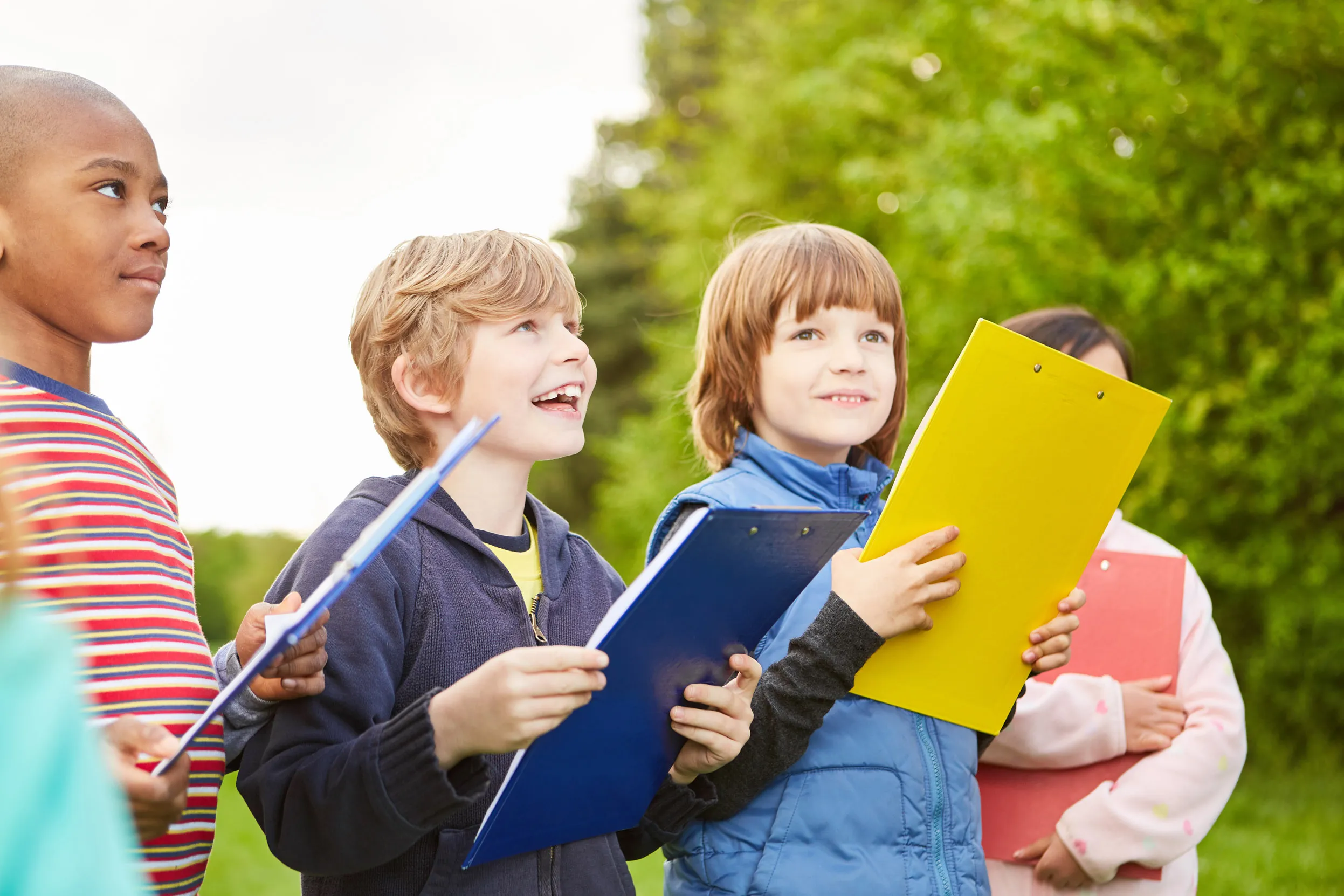 A group of four children holding blue, yellow and red clipboards learning in the woods.