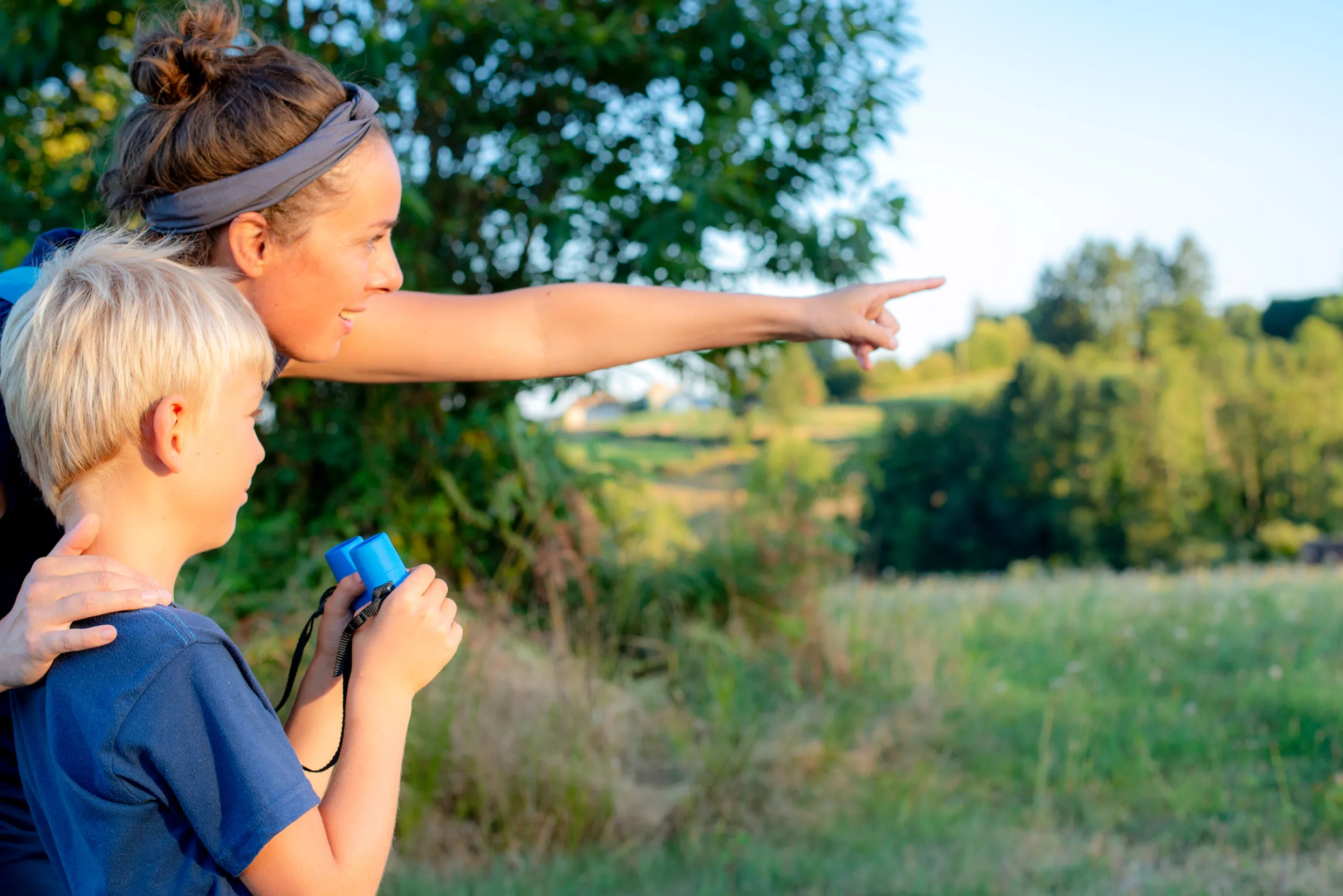 An adult pointing across a grassy field, teaching a child how to use binoculars.