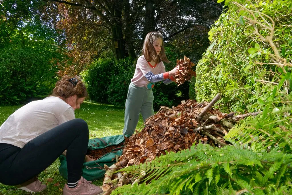A child and an adult collecting dried leaves to add to a pile of other brown garden waste.
