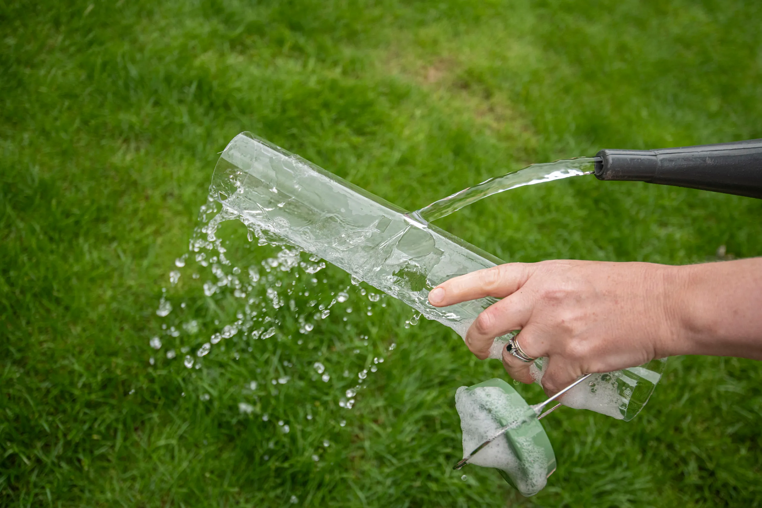 The view of a persons hand rinsing their bird feeder with a watering can over grass.