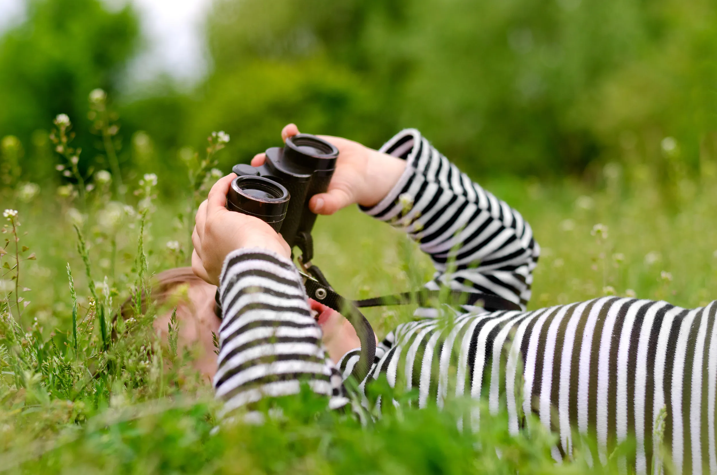 A child lay on their back in a meadow, looking at the sky with binoculars.