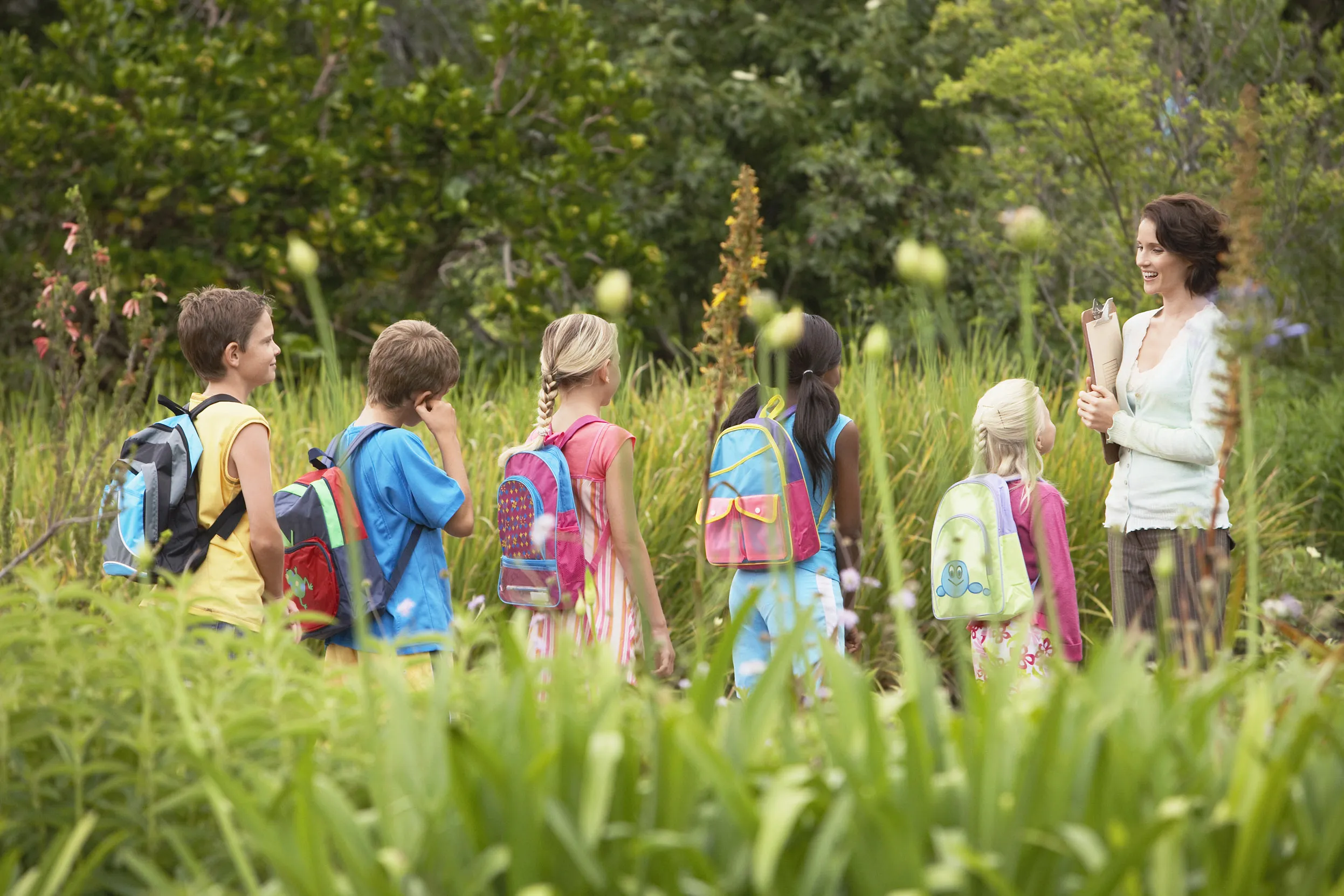 A group of school children being lead through a long grass meadow by their teacher.