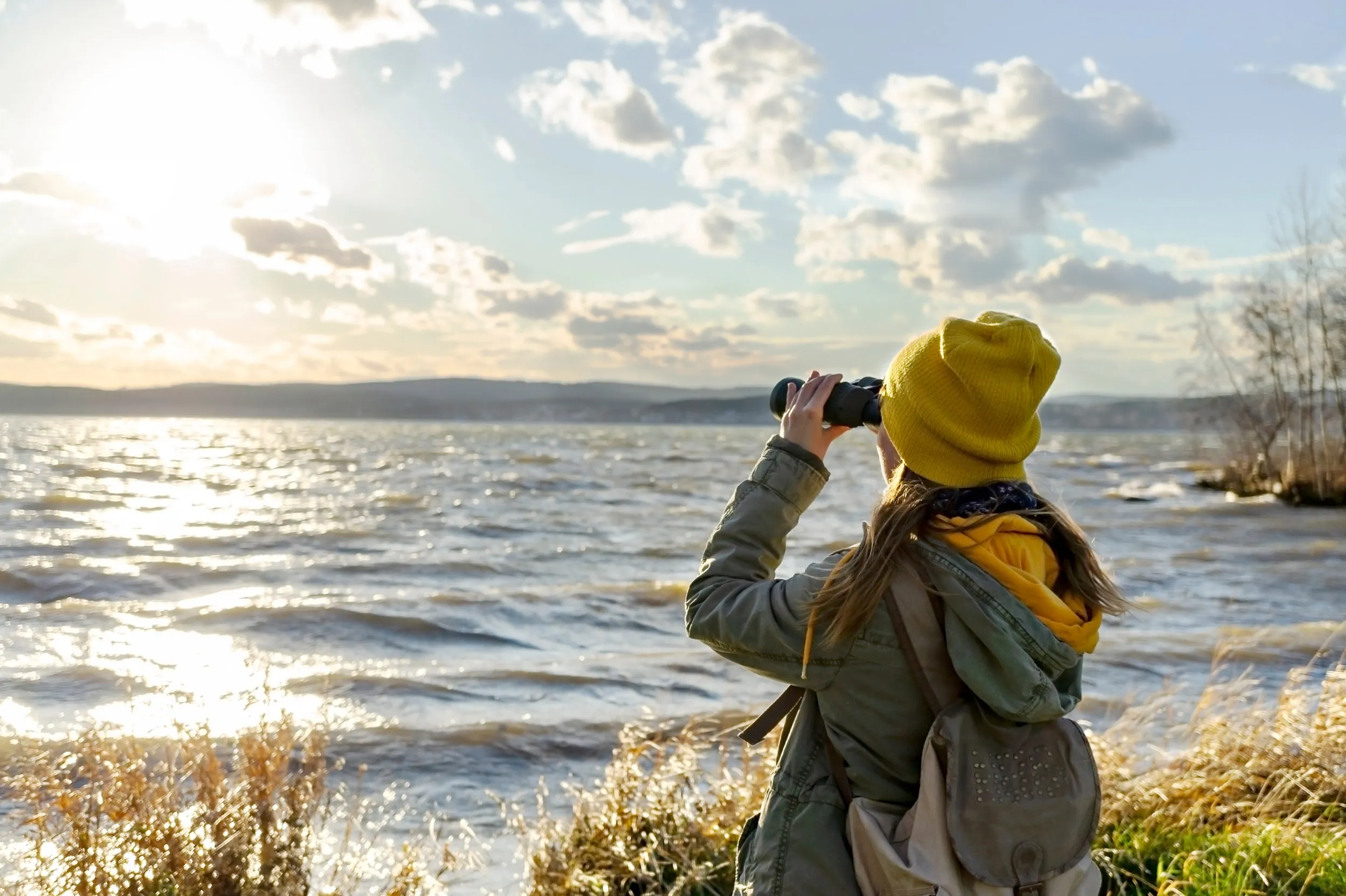 A lone women birdwatching, looking through binoculars looking over water.