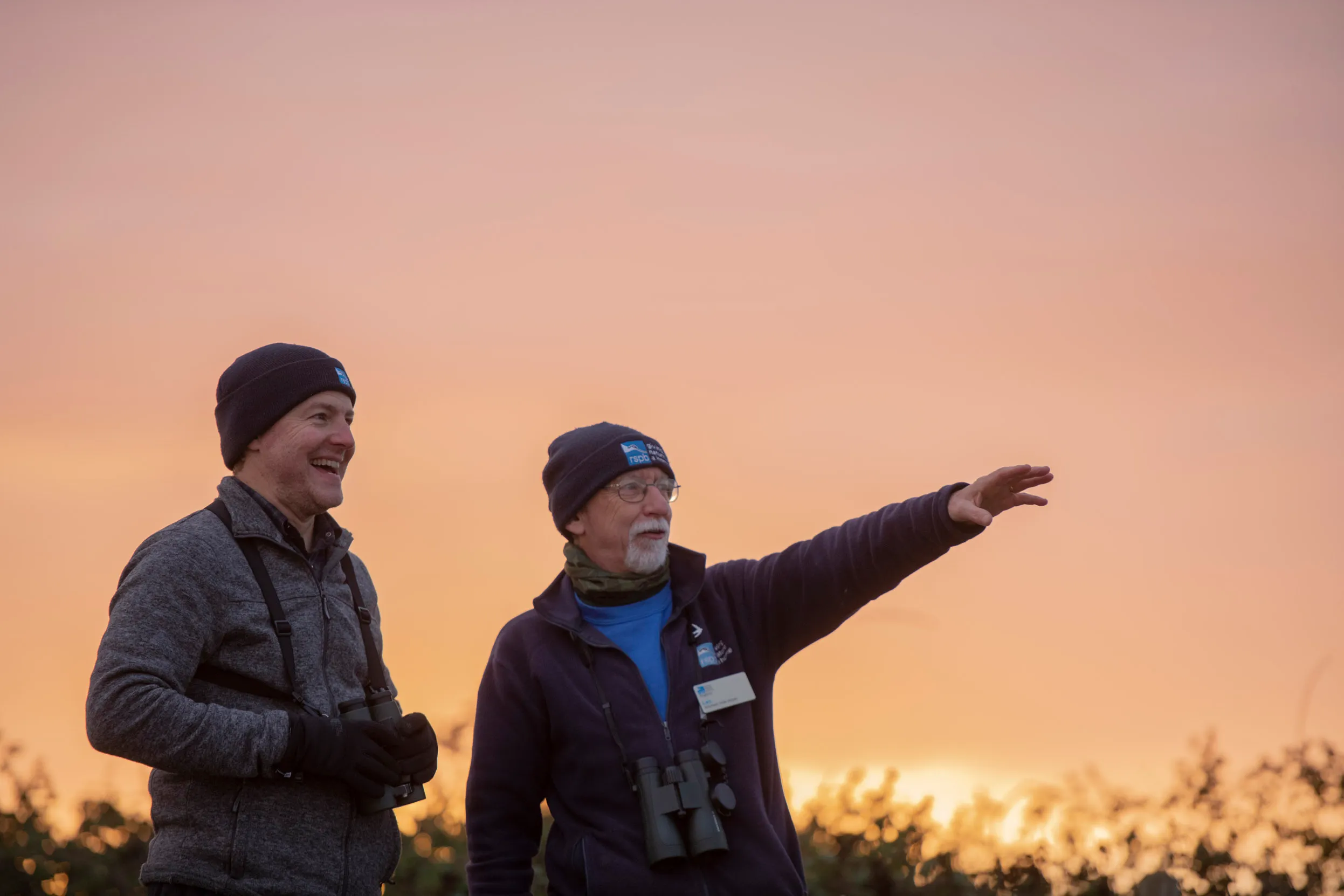 A volunteer pointing into the distance with RSPB ambassador Sam West, both with binoculars at sunset.