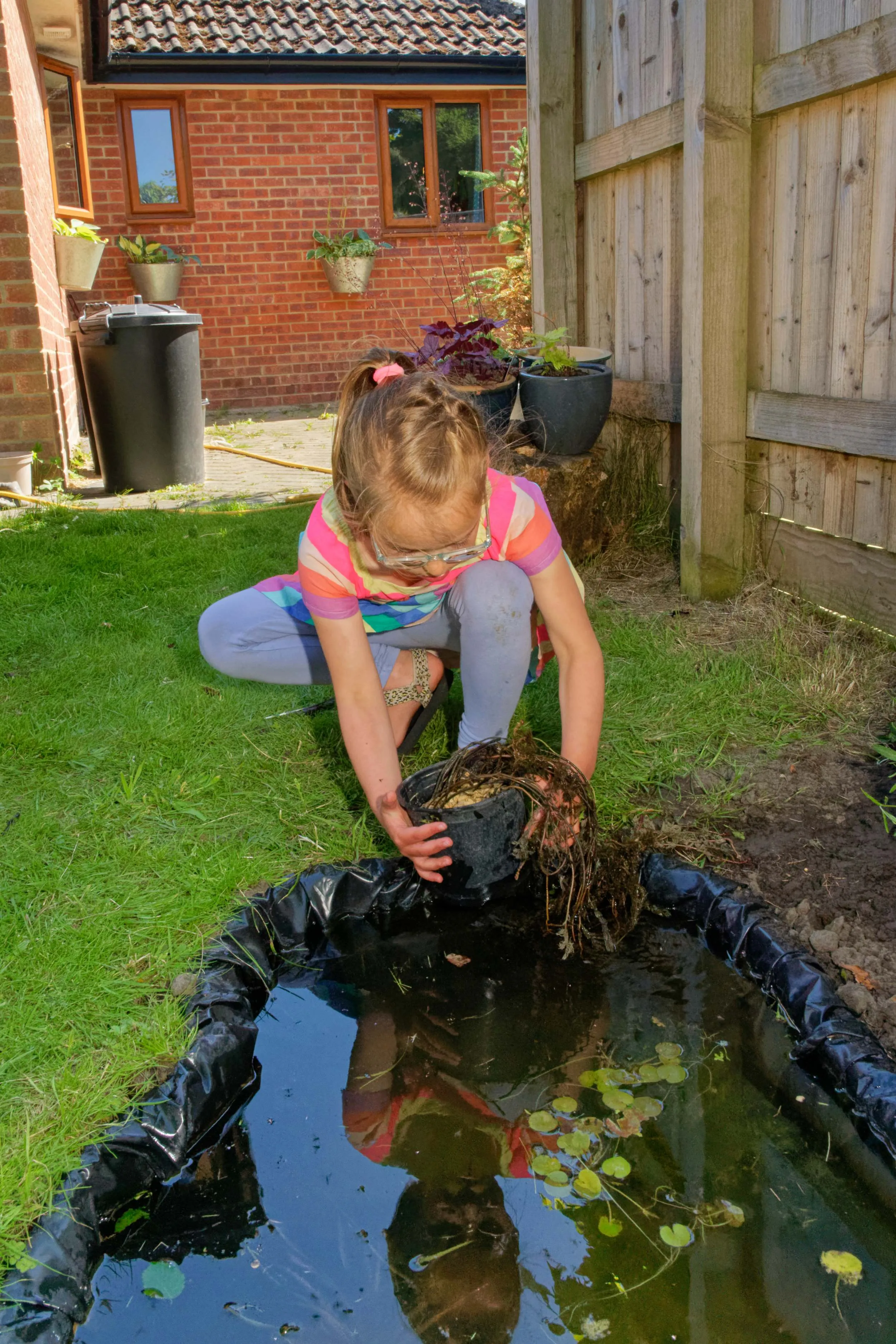 A child placing a plant pot into the water at the edge of a wildlife pond.