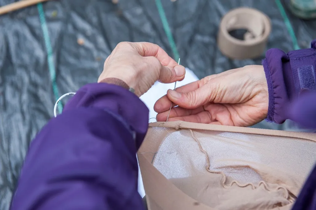 Seen from above, a person's hands sewing a pair of tights to secure them around a frame made of a coat hanger.