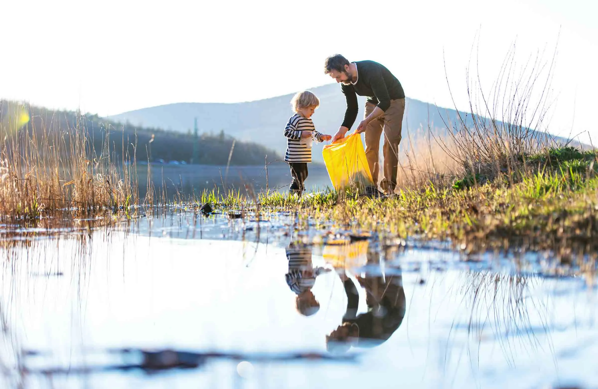 An adult and a child, stood on the bank of a lake, as the adult holds open a yellow bin bag and the child puts something inside it.