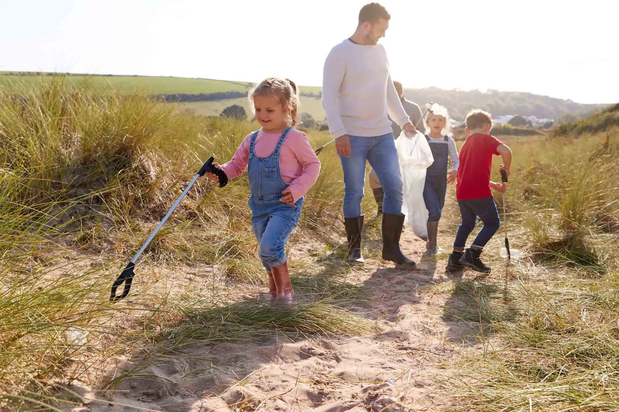 One adult and three young children on a sand dune, litter picking with a clear binbag and litter pickers.