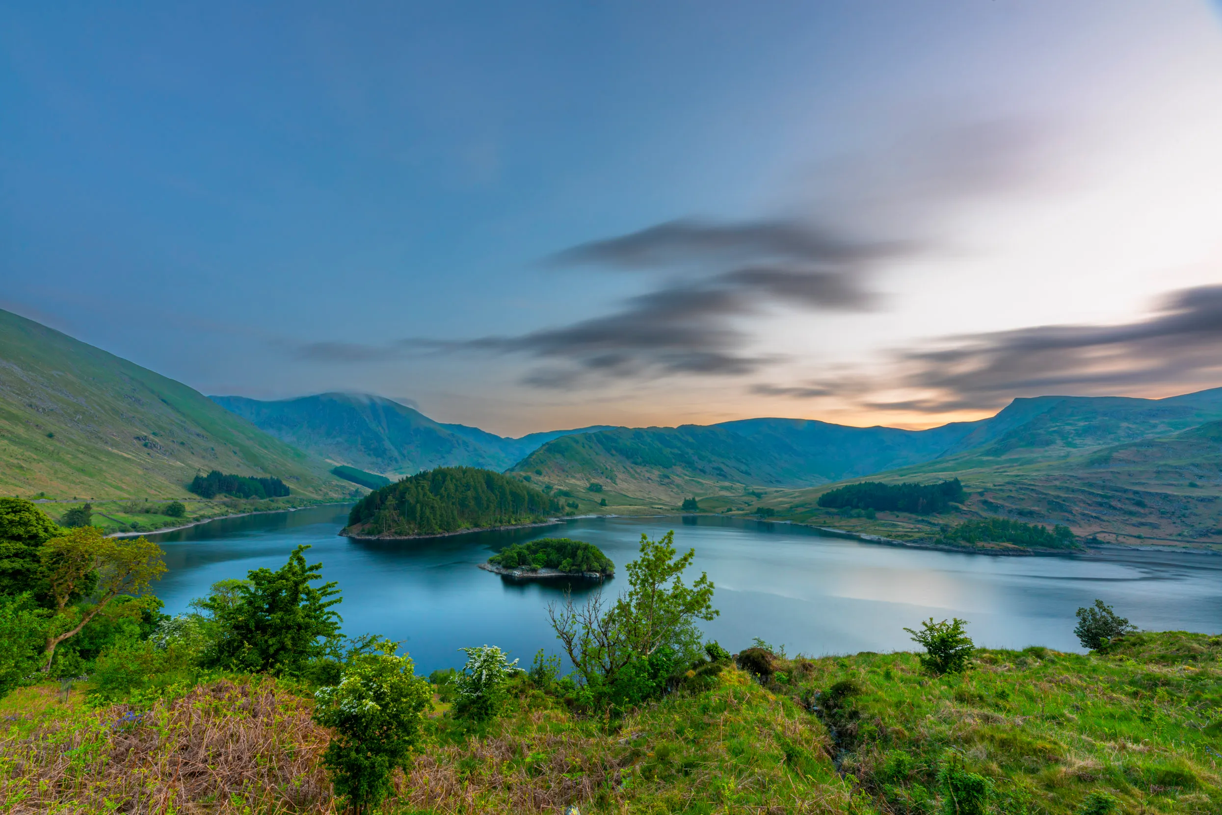 The sun setting over the main lake at Haweswater, creating a pink, orange tinge to the sky over grassy hills and islands that stand in the middle of the water.