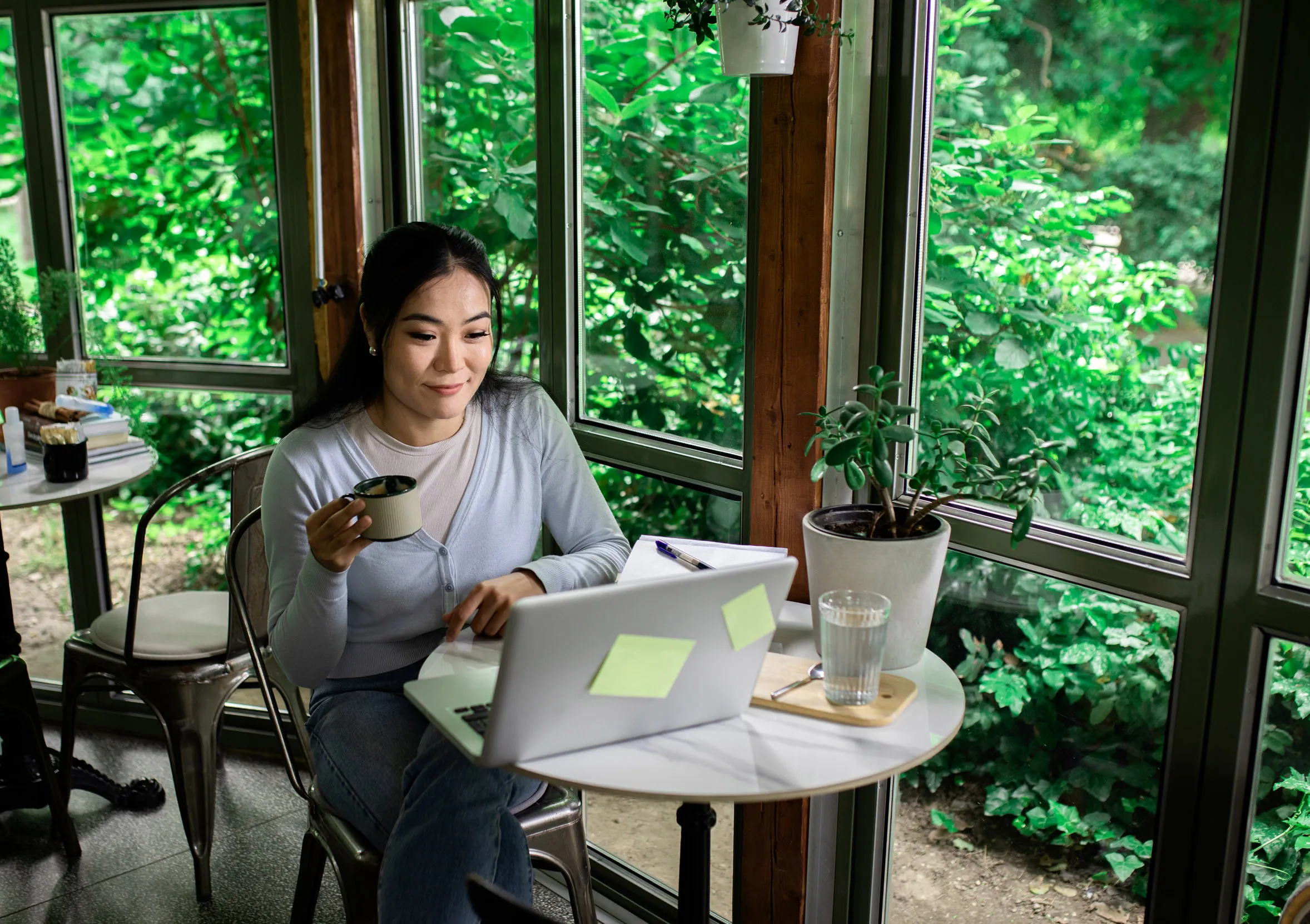 A person working at their laptop in a café, with large windows surrounded by dense greenery behind them.