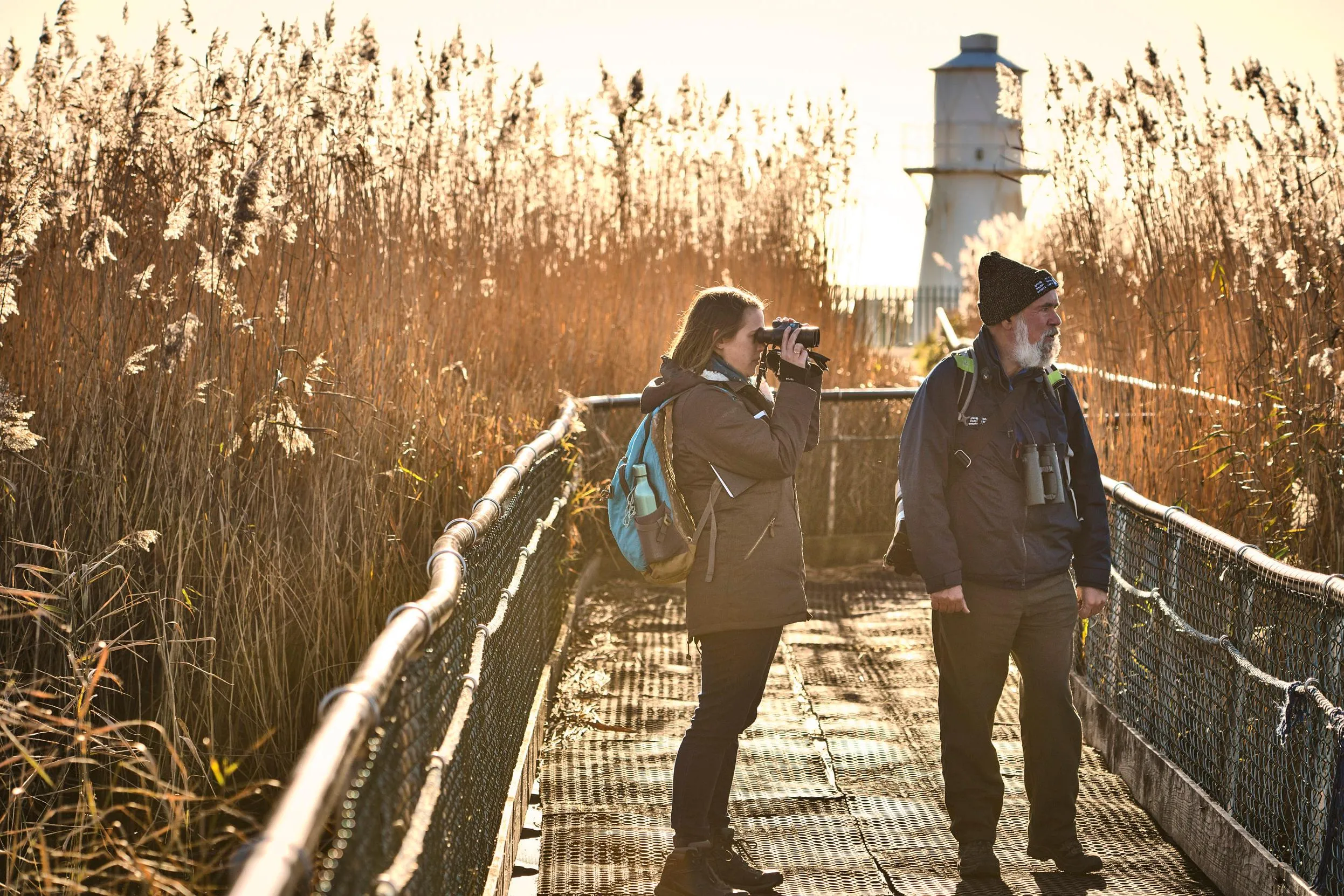 Two volunteers stood on a bridge surrounded by reeds and a lighthouse in the background.