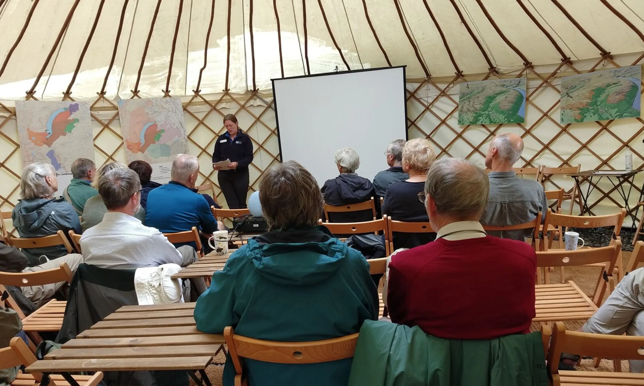 A group of people sitting down on wooden chairs, in a yurt, listening to a presentation.