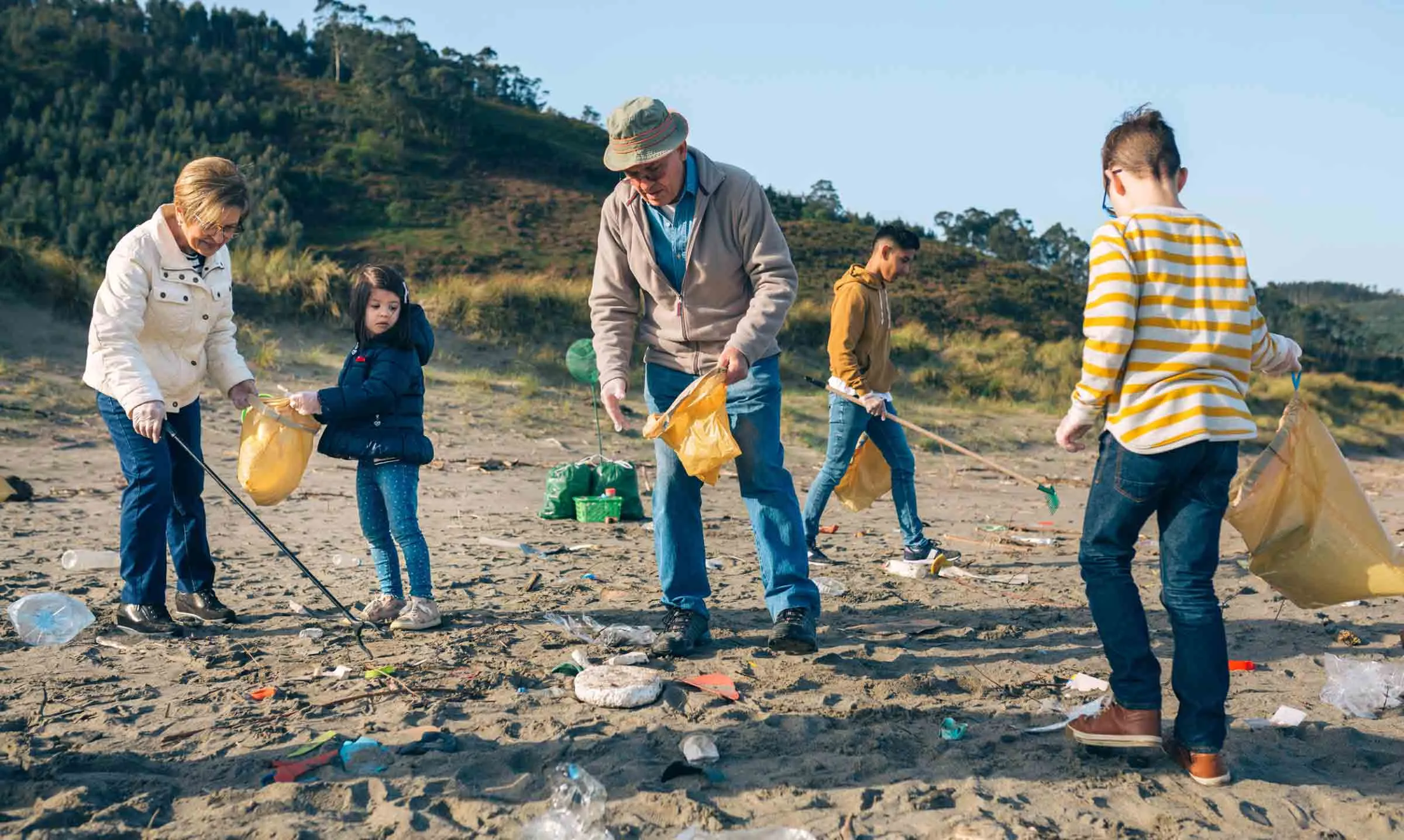 Two older adults and three children on a sandy beach, using litter pickers and bin bags to collect the plastic waste that's washed up onto the beach.