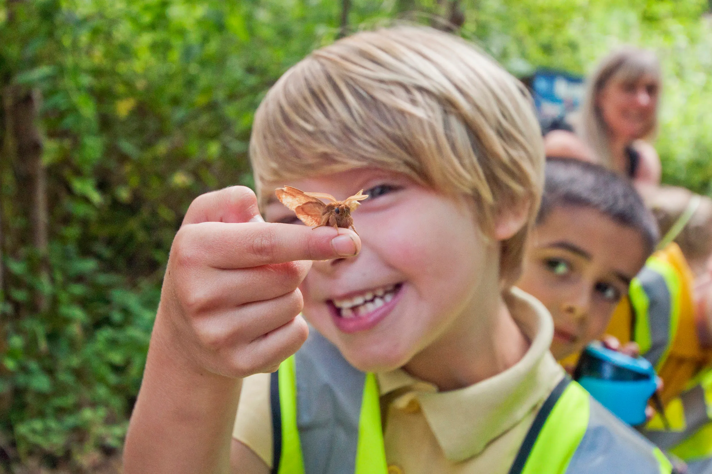 A child holding a moth in front of their face whilst wearing a yellow hi-vis jacket.