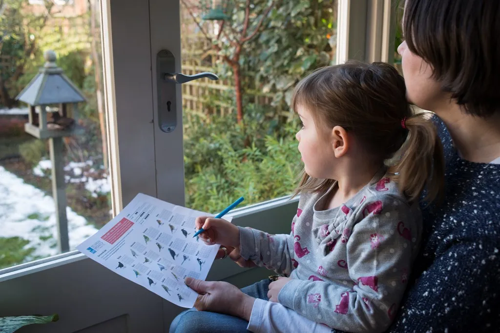 A child sat on their parent's lap, looking out of French garden doors and filling in a Big Garden Birdwatch activity sheet.