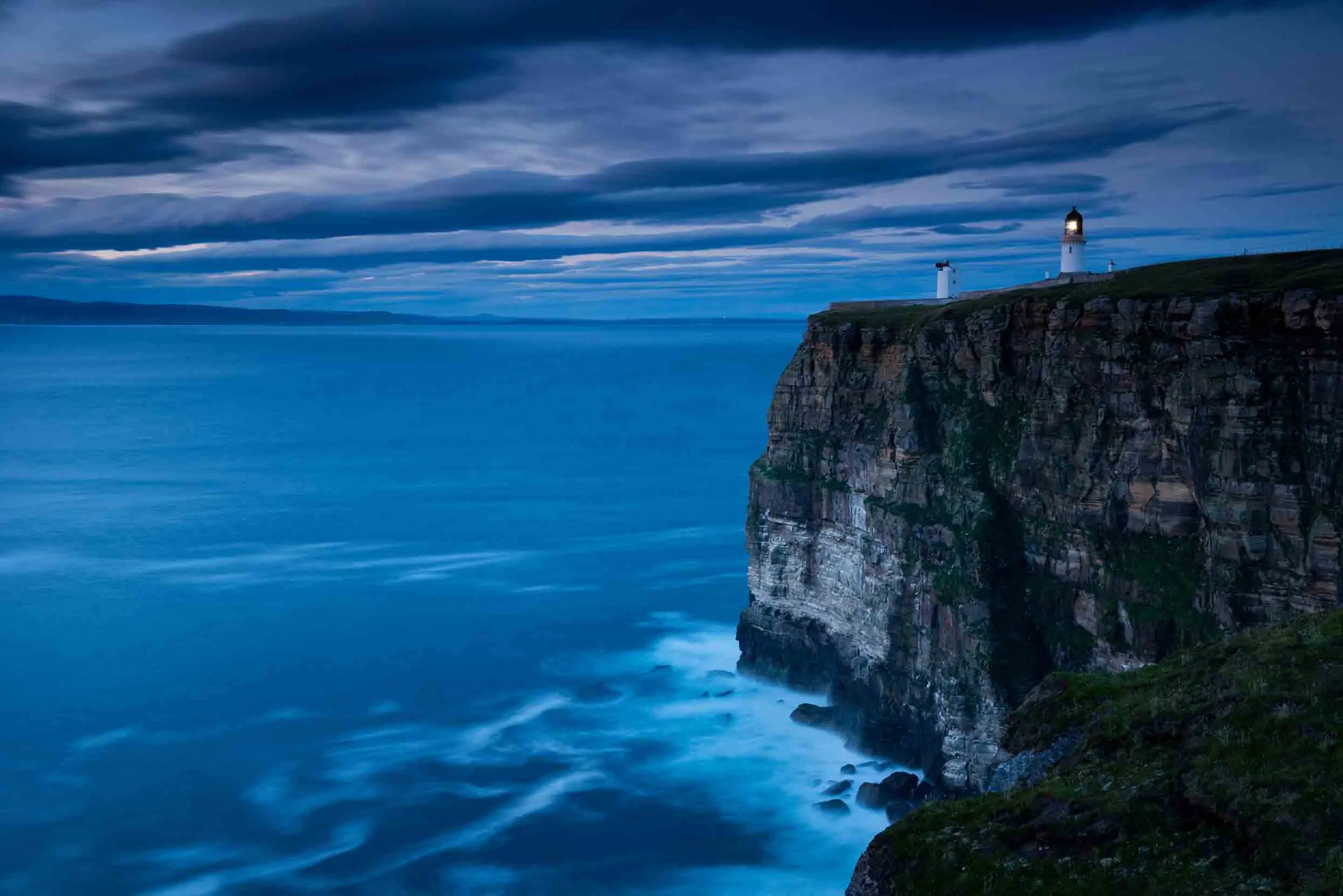 A dark blue sky looms over the calm blue sea at the base of the Dunnet Head cliffs, the light house can be seen at the top.