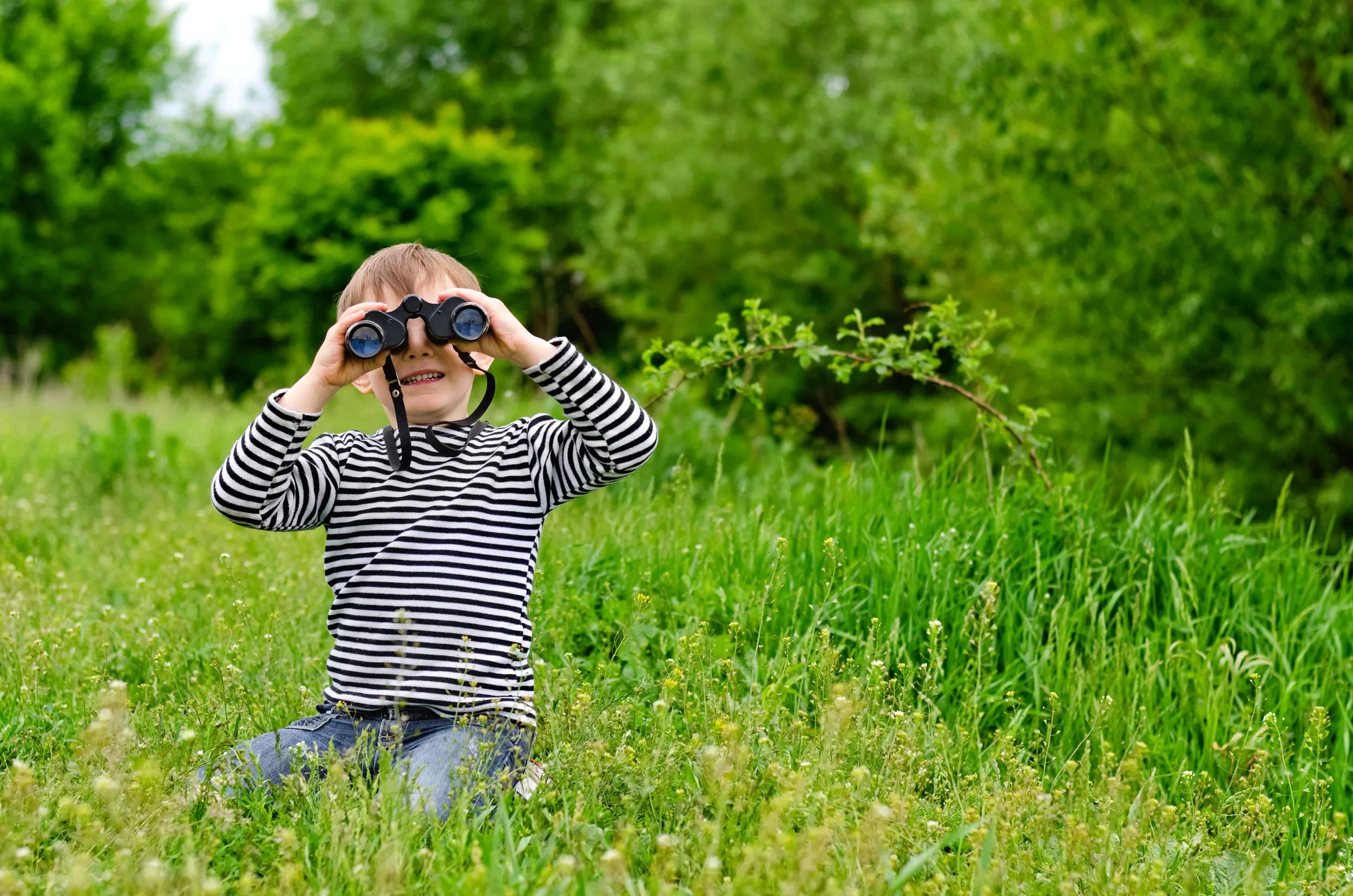 A child kneeling in a meadow, looking through binoculars towards the camera.