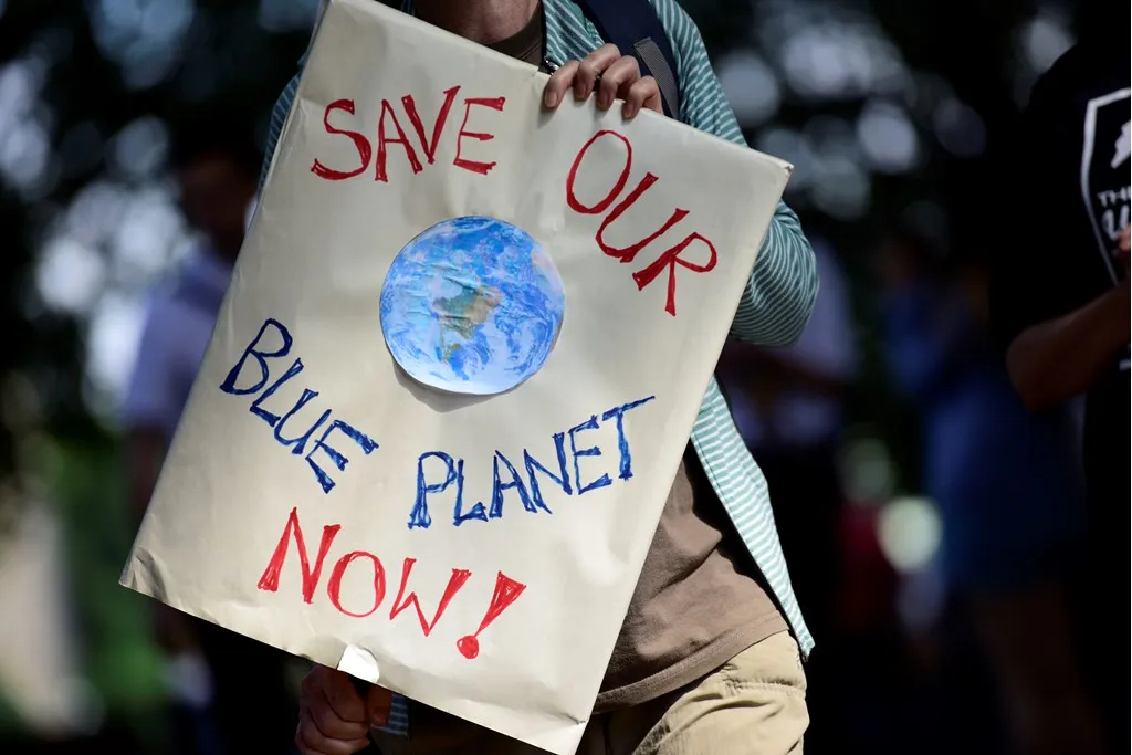 A person holding a paper sign which reads "Save Our Blue Planet Now!", with a picture of the globe in the centre.