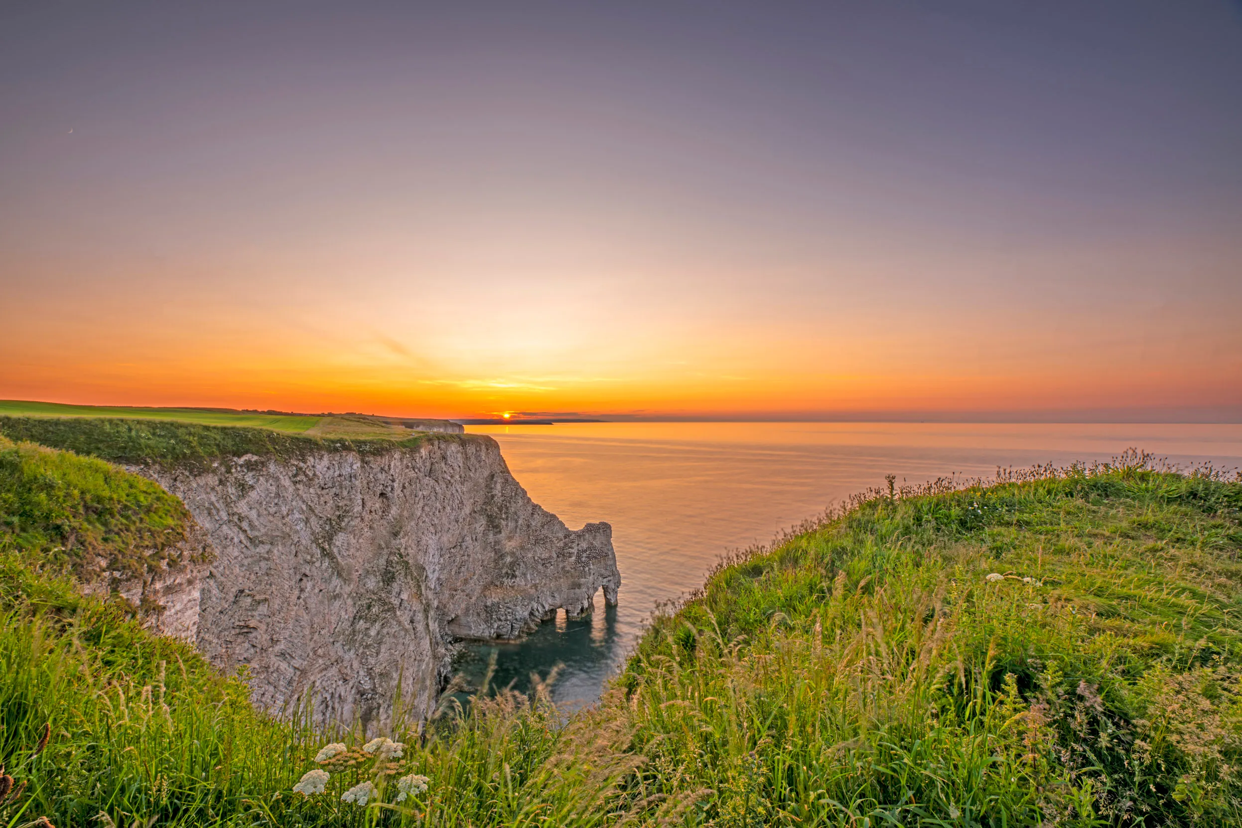 The view of the cliffs at Bempton overlooking the water at sunset, showing surrounding grassland and the arch rock formation in the cliffs.