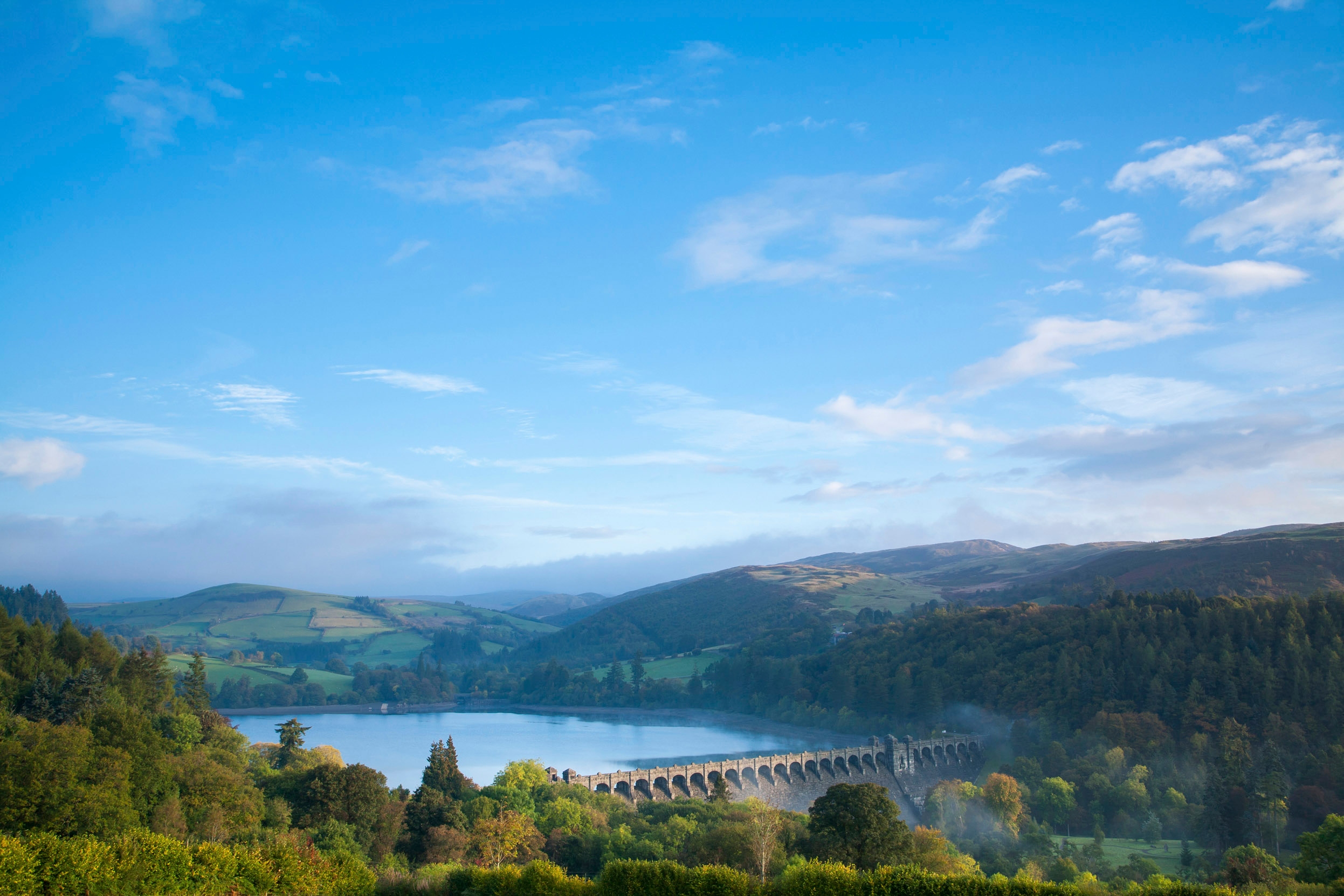 A view of Lake Vyrnwy and the stone wall, surrounded by trees and a blue sky with wispy white clouds.